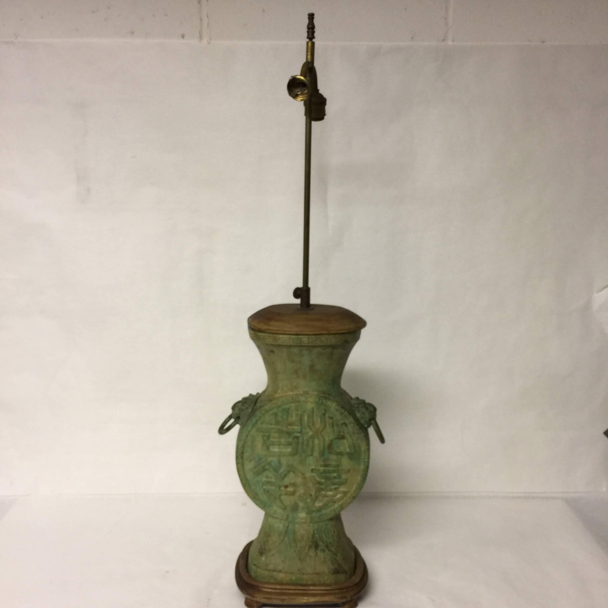 In the style of Frederick Cooper, this Asian Style, antique patina table lamp has a Greek key design, characters, lotus leaves. There are lion heads with rings in their mouths on either side. The lamp sits atop a wooden base and has a wooden top.