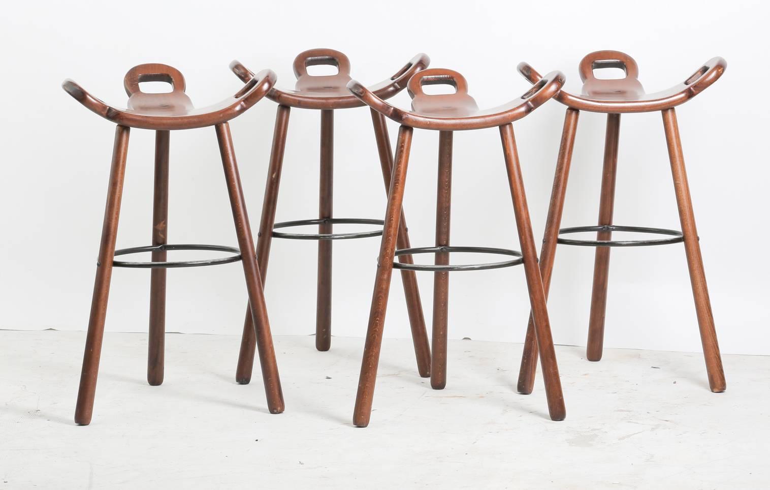 Set of four "Marbella" Brutalist dark-brown stained beech bar stools, each with three legs and black-lacquered metal rest ring. Manufactured by Confonorm, 1970s. Seating has odd curved T-shape with three handles. The handles are not only