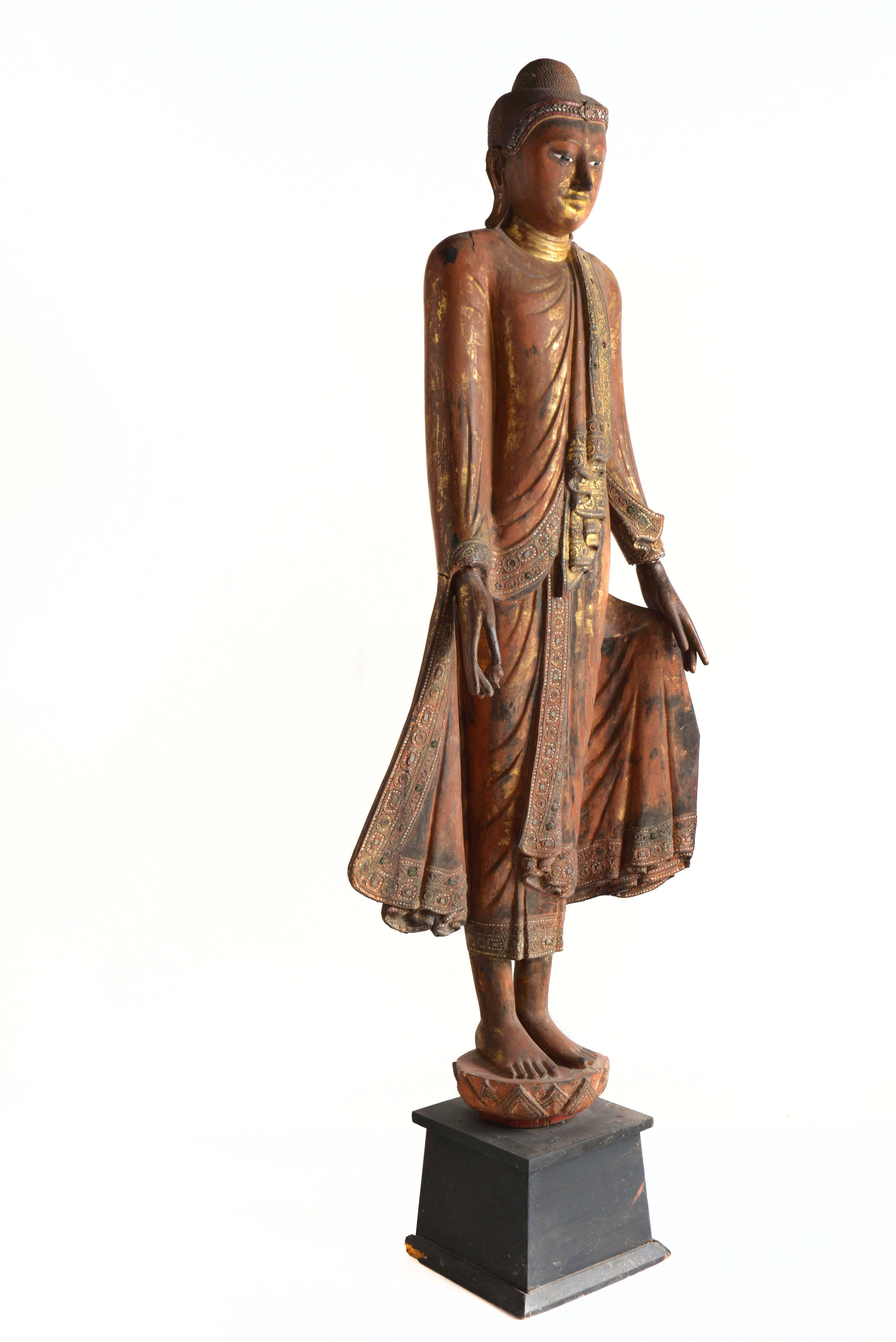 Standing Mandalay Buddha with flowing robes, holds both hands in the Varada gesture symbolizing fulfillment of all wishes, boon and charity. The Buddha's right hand is holding a myloban fruit offering and he stands on a Lotus throne. Wood with
