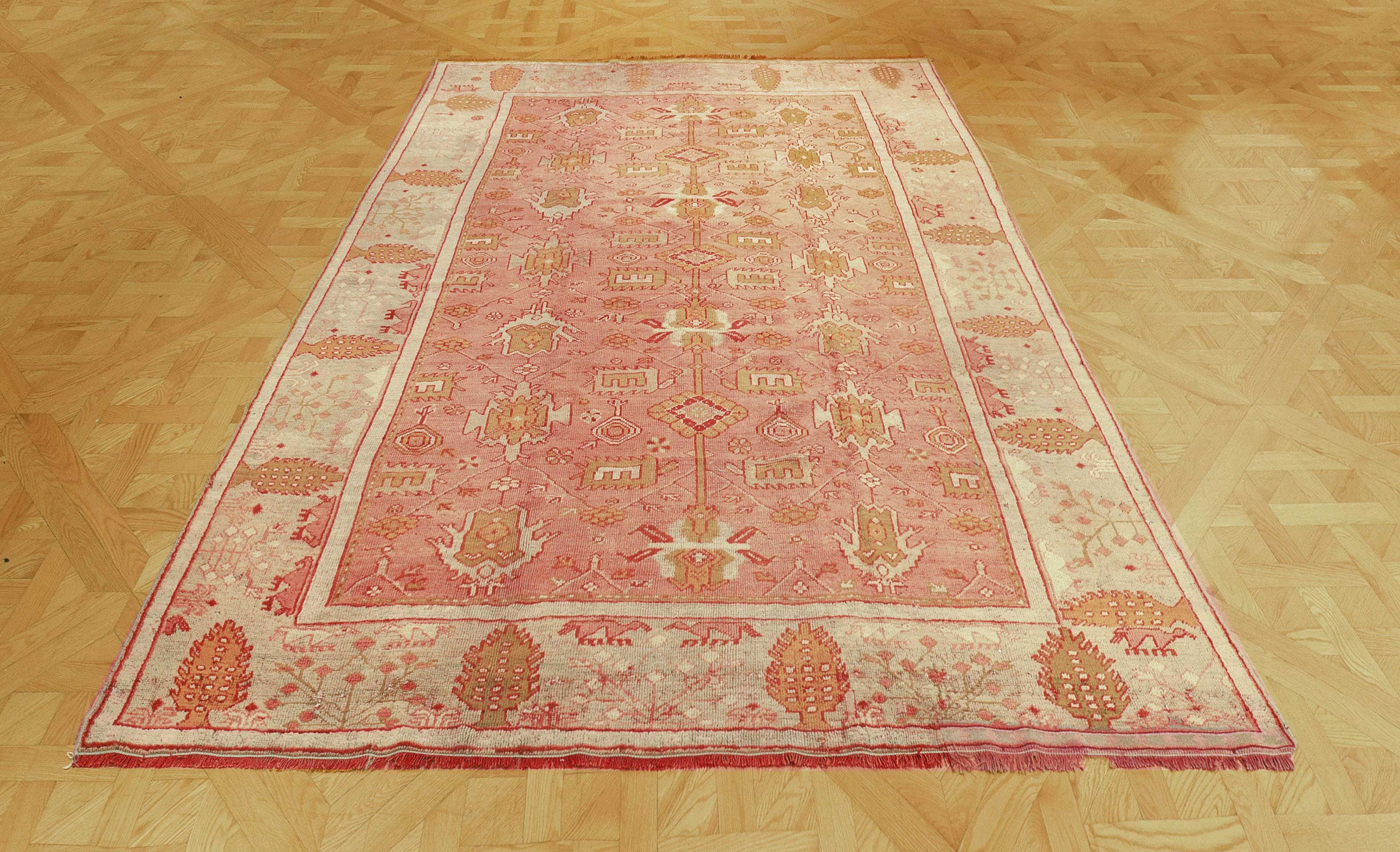 The terracotta red field with an overall angular shaded green and ivory palmette and floral vine, in an eau-de-nil border with stylised cypress trees alternating with a delicate floral sprays between plain ivory stripes.

Oushak (or Ushak) rugs