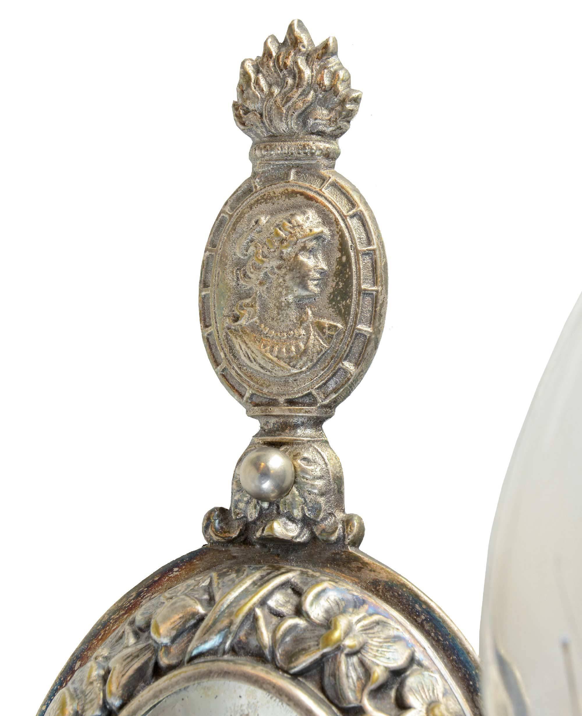 These decorative silver over cast brass sconces, manufactured by Moran & Hastings of Chicago, IL, feature a decorative floral border around a beveled mirror on the backplate, with the same leafy pattern around the bobeche, as well as a cameo of a
