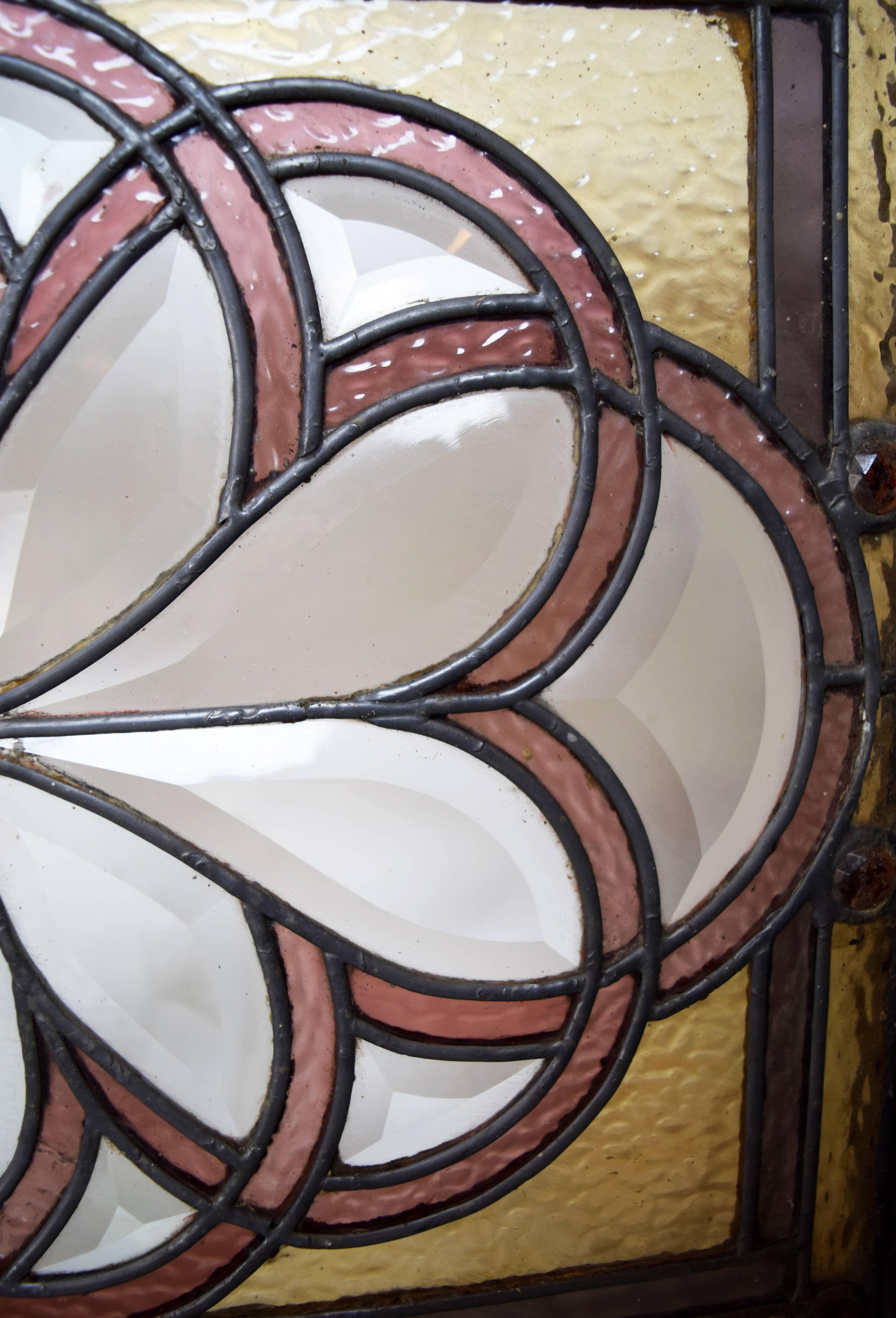 This wonderful, unique curved Victorian window features bevelled glass squares in its centre, beautiful orange jewels lining the top and bottom on a yellow glass border, and two symmetrical curving floral dark pink glass designs on the sides. This
