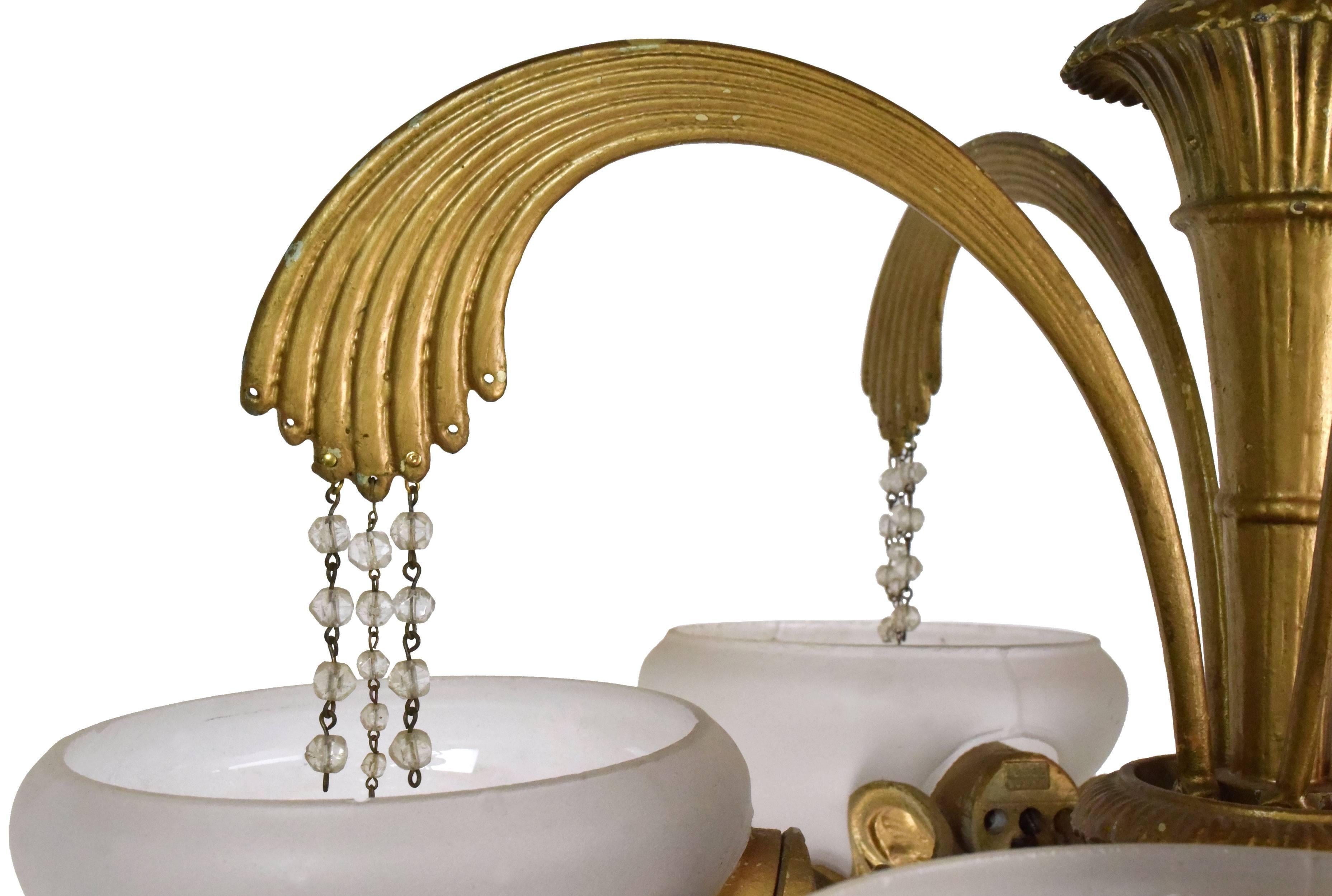 This Classic Art Deco brass chandelier features five opaque shades and gracefully curving arms. Small crystals hang into the bowls from each arm, creating a cascading, water-like effect. The bottom of the fixture is equally beautiful, where