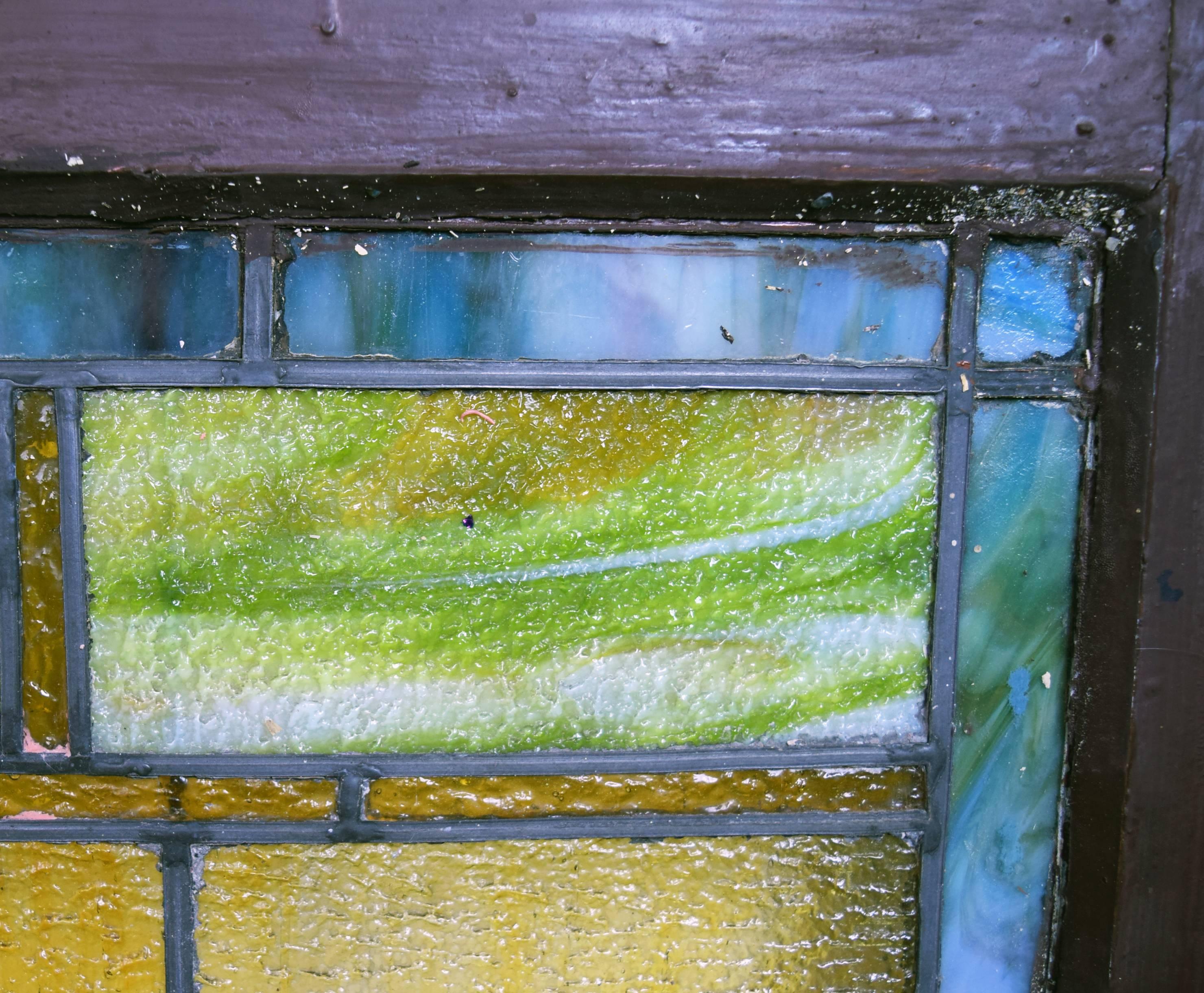 This beautiful Arts & Crafts window features a wonderfully detailed yet subtle hand painted rondelle, bright yellow textured glass panels, slag glass green, blue, and multicolored smaller panels and deep green leaf designs surrounding the rondelle.
