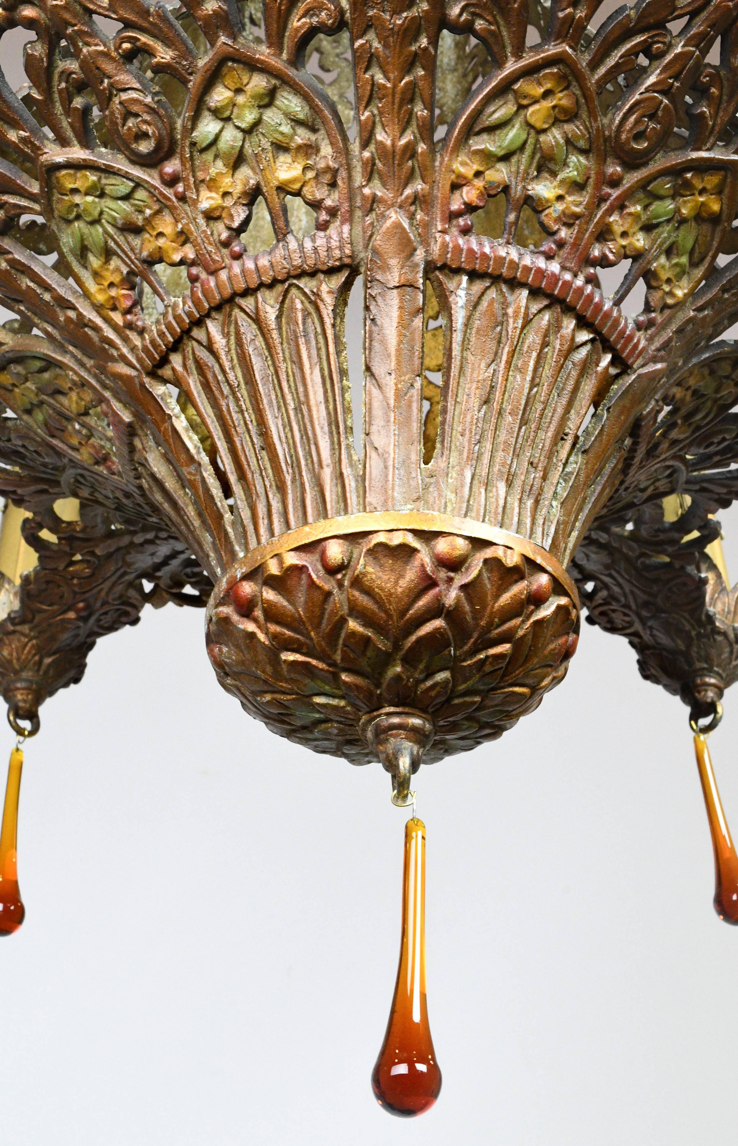 This gorgeous cast aluminium chandelier features intricate filigree details decorated in a polychrome finish of many colors. The leafy candle holders align with the botanical details throughout the fixture's body, and amber teardrop crystals add an