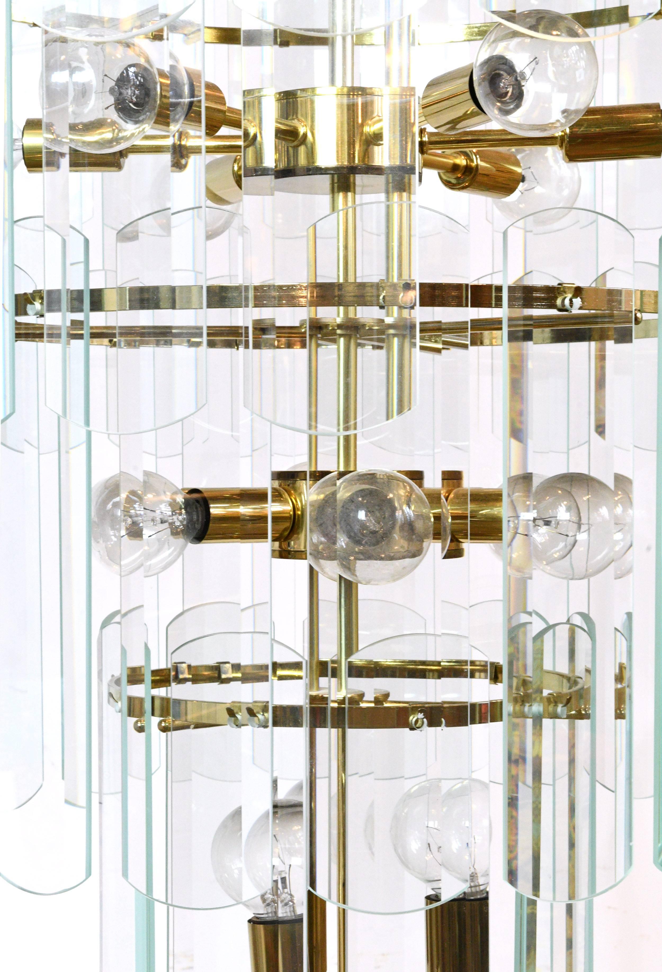 This large chandelier features a swirling brass body surrounded by 93 glass panels spread out between four-tiers. The blue-tinted panels offer clean lines and crisp reflections. 27 bulbs give the fixture a dazzling radiance that will light up any