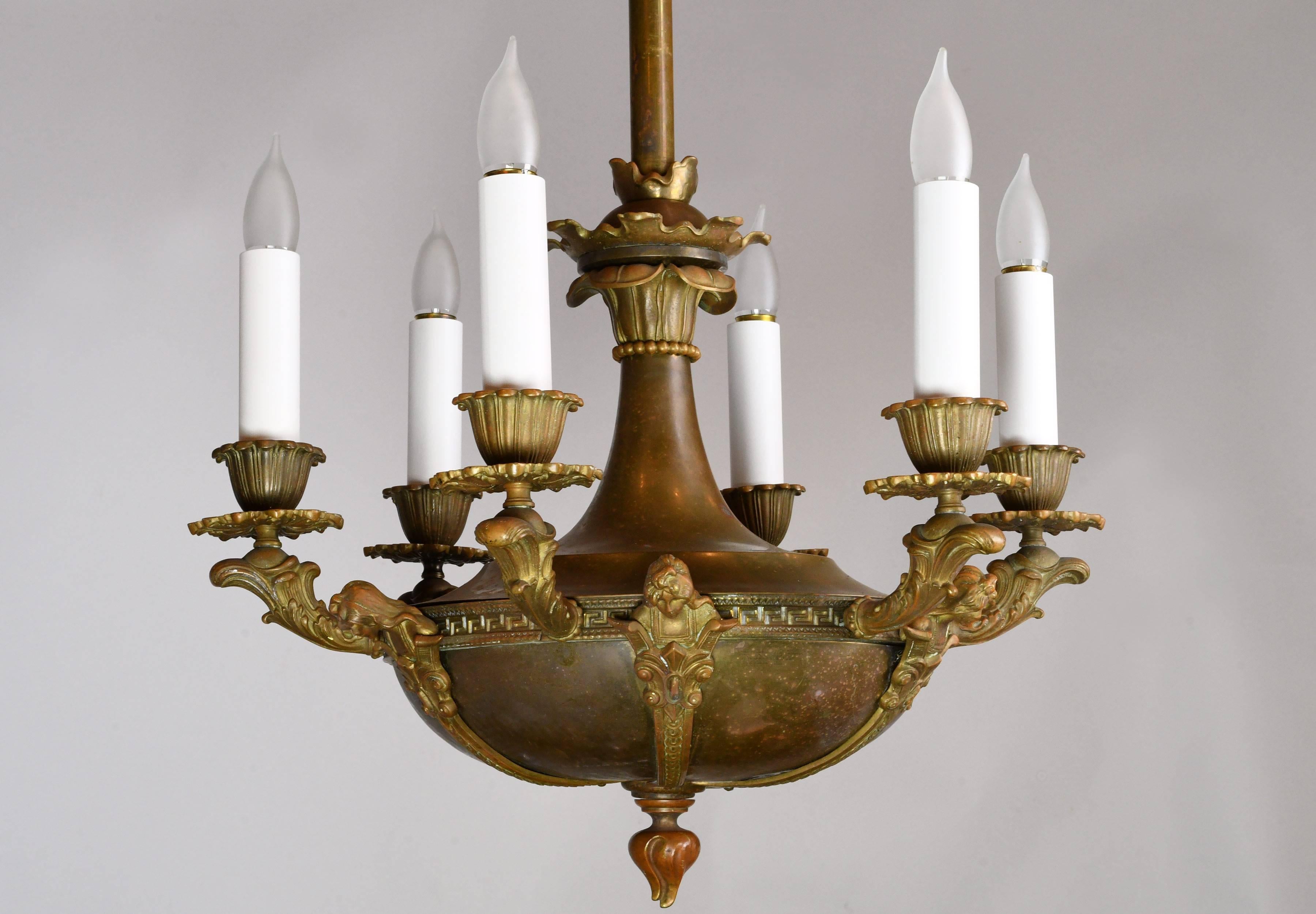 Crafted out of bronze, this six candle fixture has aged beautifully and has small amounts of varying colored patina throughout. The candles of this intricate chandelier are interspersed with human figures, reminiscent of figureheads on a mighty