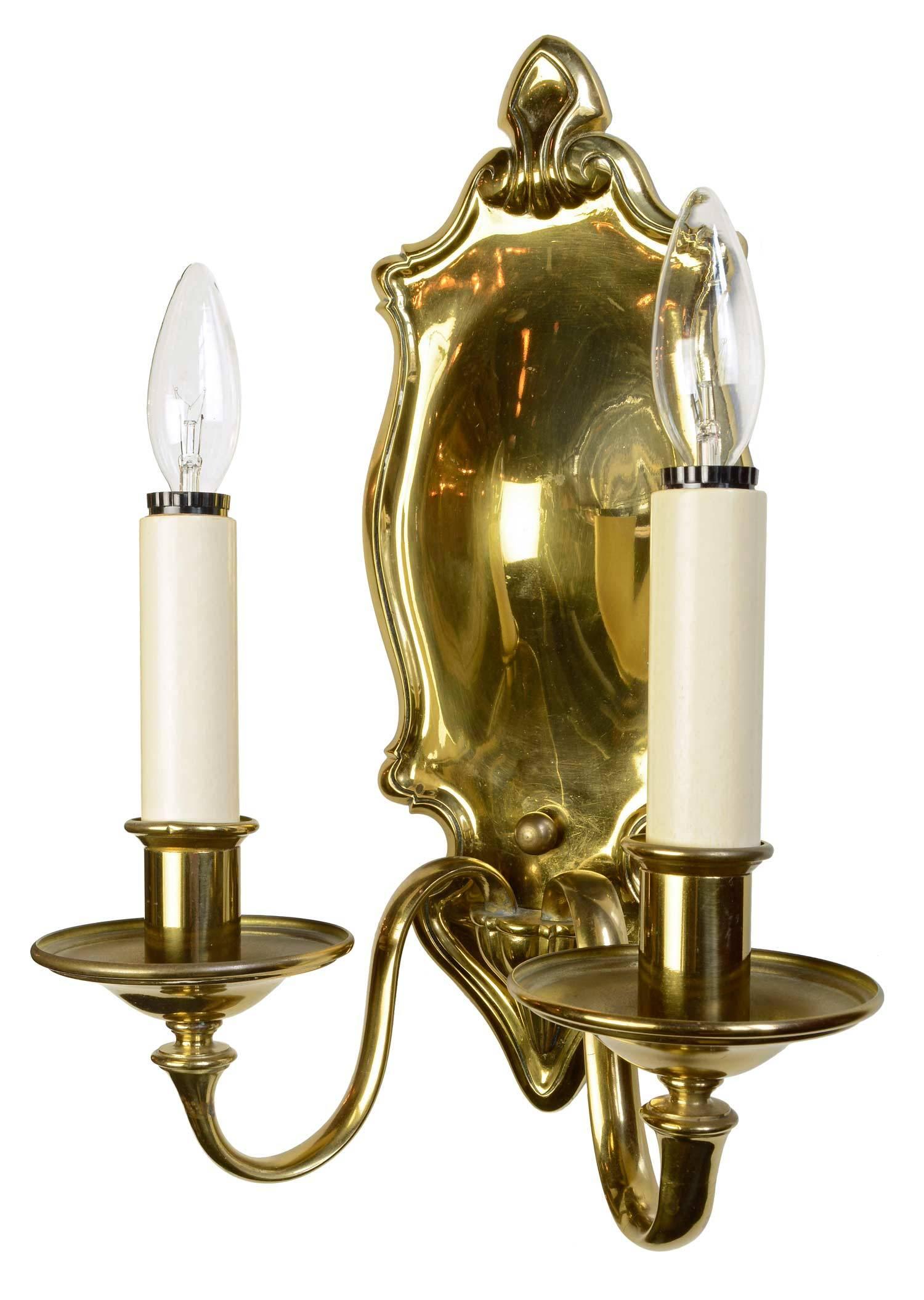 This pair of oversized cast brass two-arm Colonial Revival sconces, created circa 1920s, are signed Bradley & Hubbard, a company known for its quality of craftsmanship. The simplicity of design and convex shaped backplate reflects light from its