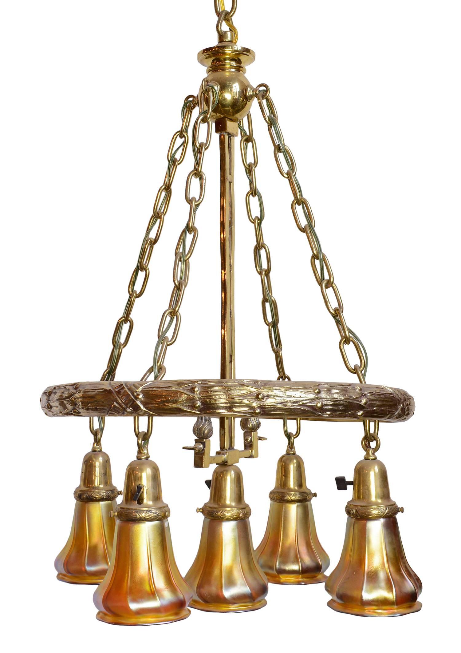 This finely restored and polished chandelier features the laurel leaf pattern that decorates the large brass ring and the bell shaped shade holders.  The center rod is the original gas/electric mechanism and the five aurene shades are signed