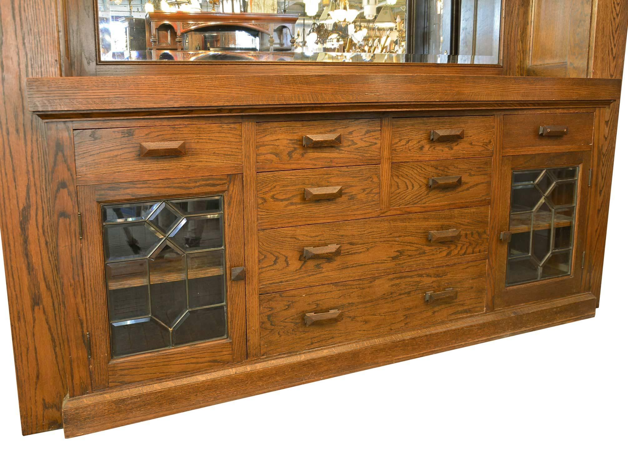 Built-in oak buffet with beveled mirror, leaded beveled windows on upper and lower cupboards, multiple drawers on bottom, and brass single-arm Sheffield sconces with art glass shades. Original oak finish and craftsman pulls. 
This unit is a built