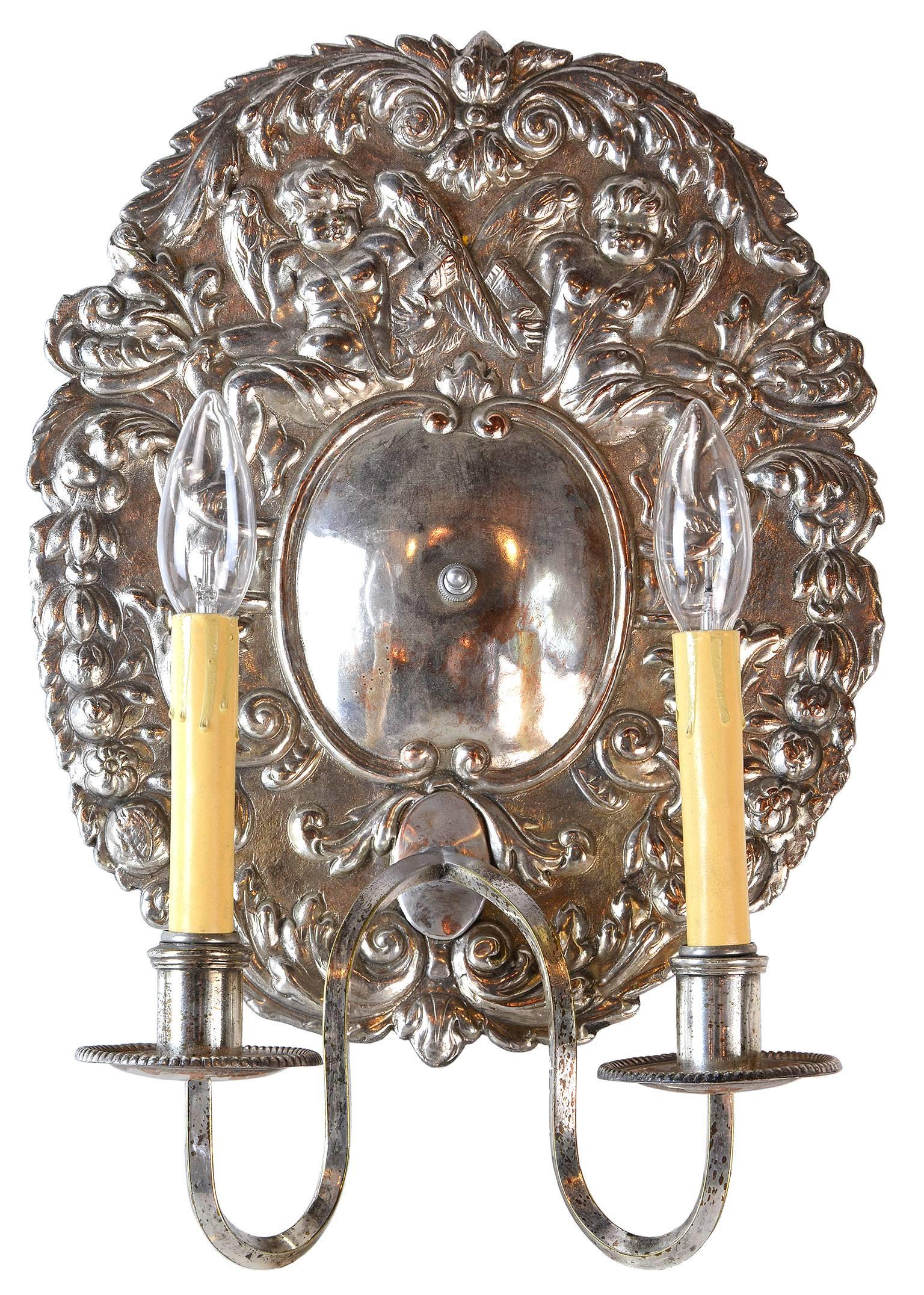 This two-arm sconce pair from circa 1880 was converted from candlesticks to electric, and has a large decorative silver backplate depicting cherubs and floral details. The making of this piece was exceptional, and its subject matter, in the Rococo