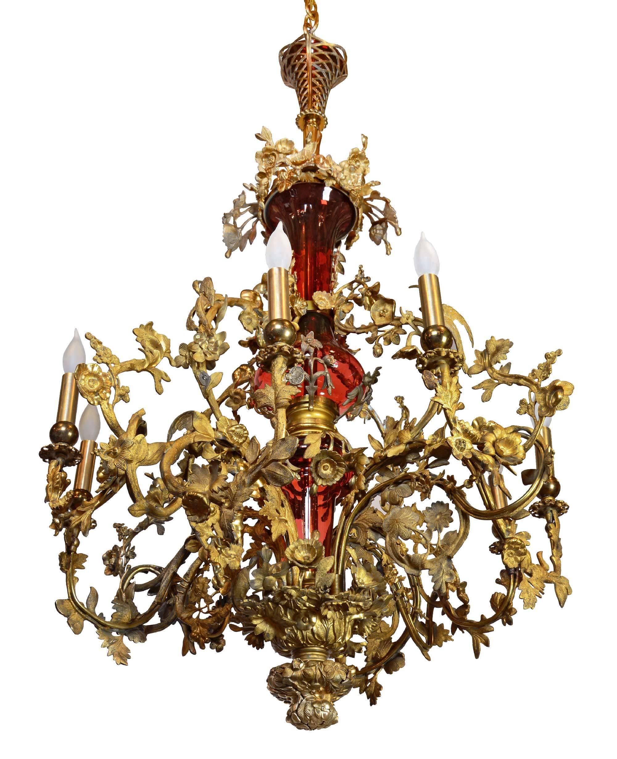 This beautiful converted gas chandelier features ornate, leafy scrolling arms coming up from a floral finial with bird details and brass flowers spilling from the top of a cranberry glass urn. Complete with brass candle covers and original matching