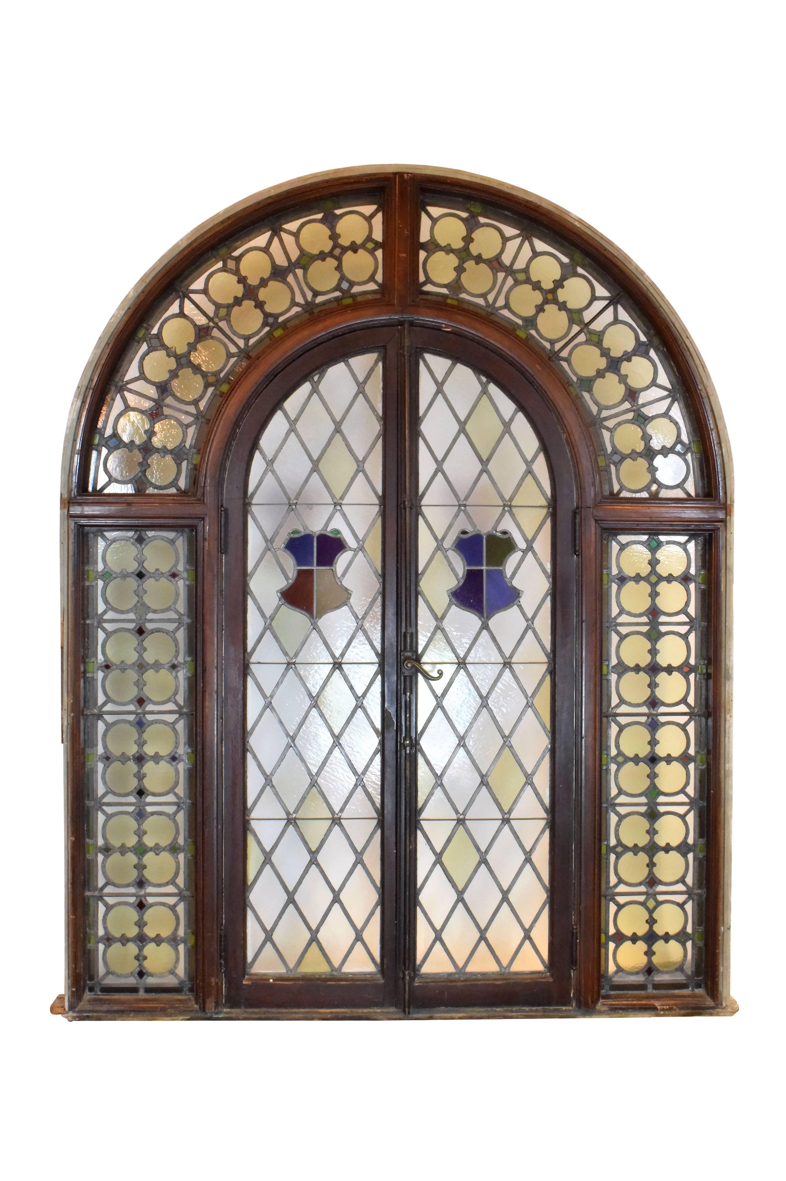 This gorgeous arched French window unit features colorful stained glass details throughout and decorative metal latticework running across the glass. A smaller window within the unit unlatches with a beautiful brass cremone bolt. Richly stained wood