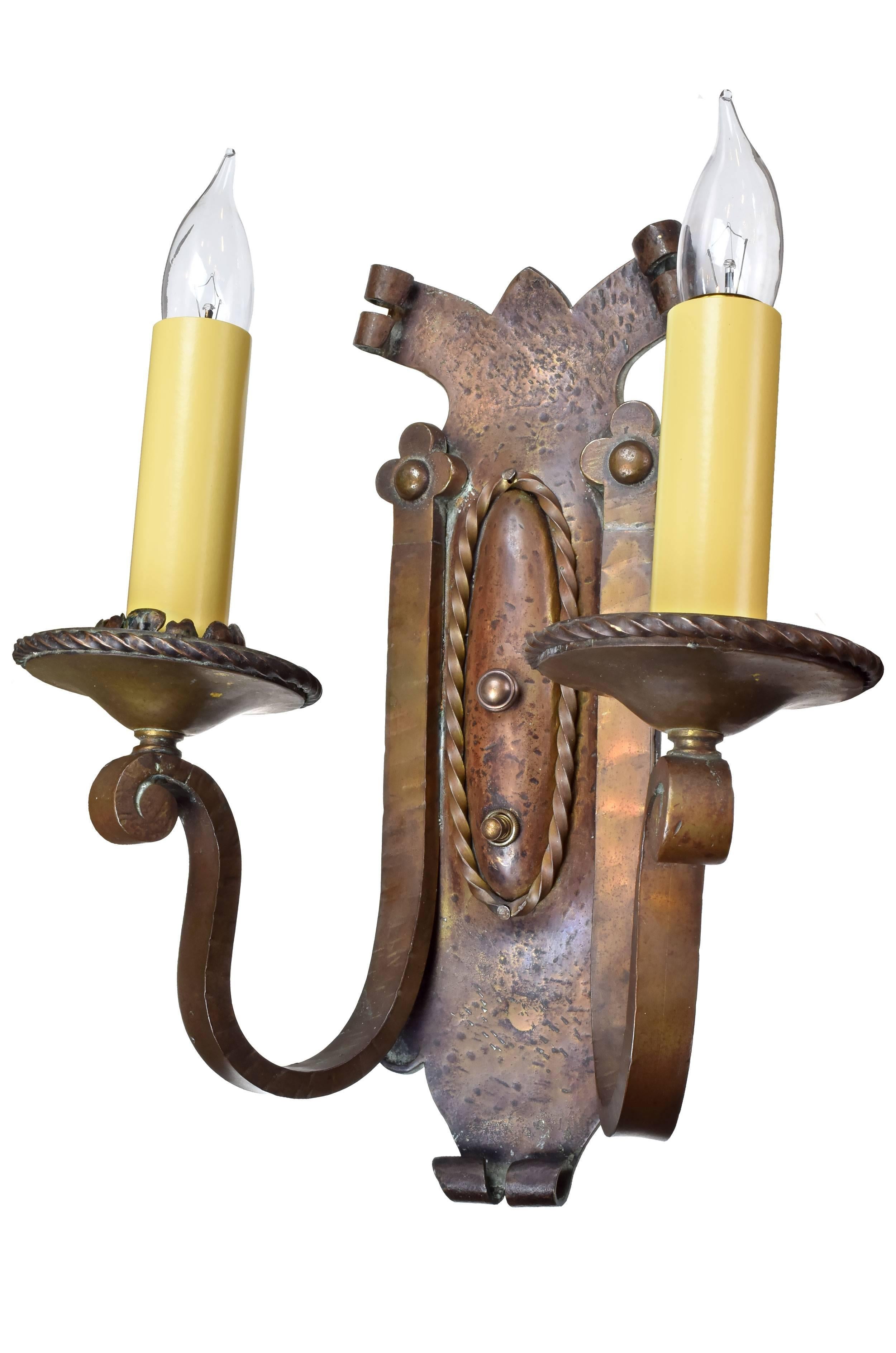 These heavy hammered brass two candle Tudor sconces feature attractive, curving lines throughout and organic ornamentation surrounding the candlesticks. These fixtures are gorgeous on their own and even more striking when grouped together,

circa