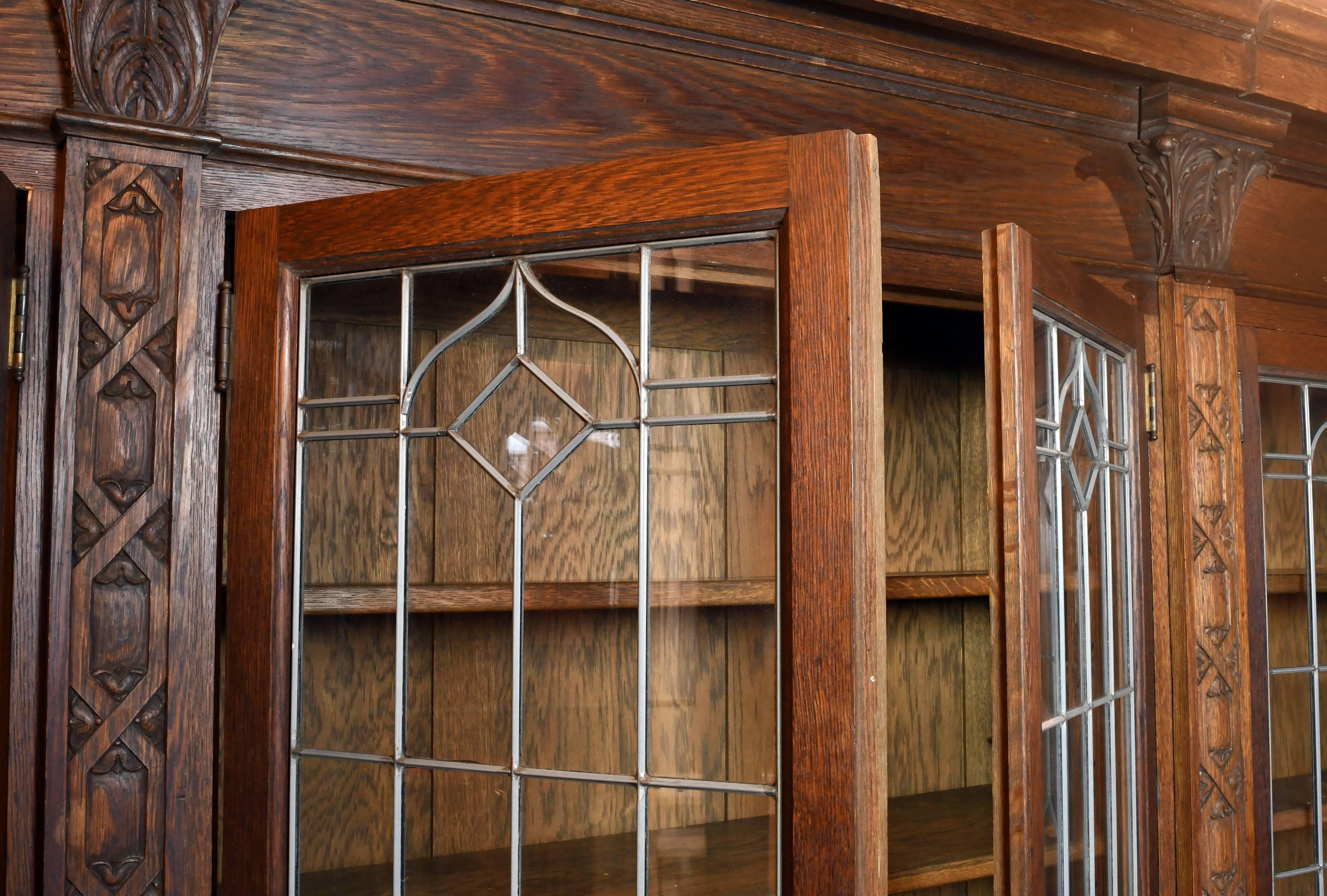 This long oak bookcase unit is complete with four sets of leaded glass double doors that each contains four shelves of adjustable height. Many details abound in the woodwork; bellflowers mixed with woven diamonds are arranged in narrow vertical