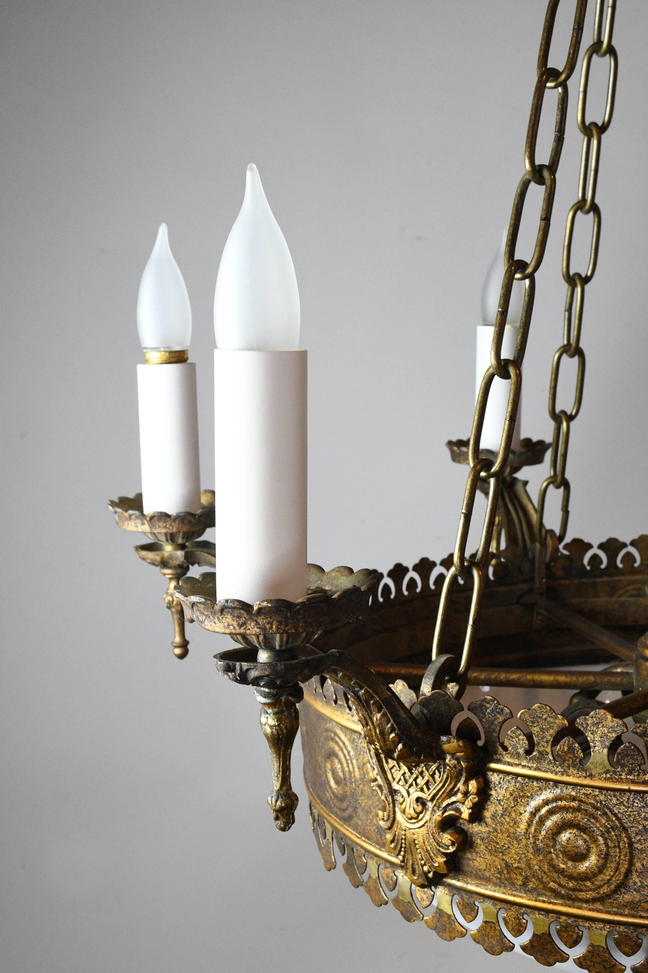 This brass chandelier has six candle holders surrounding the exterior, while three interior sockets illuminate the bent glass globe. The chandelier has Tudor motifs and intricate cutout detailing throughout. Beneath the white globe is a delicate