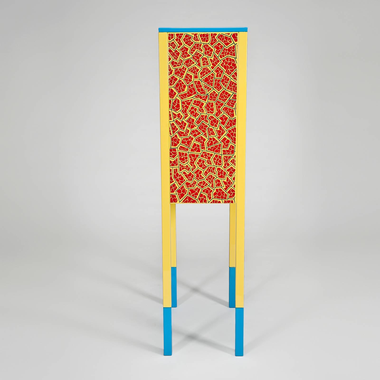 Glass front cabinet with red lacquered doors and yellow trim. The yellow legs are tipped in blue to match the blue at the top of the cabinet. The Sowden- designed pattern on the sides is stenciled in red and blue on the yellow background. The grey
