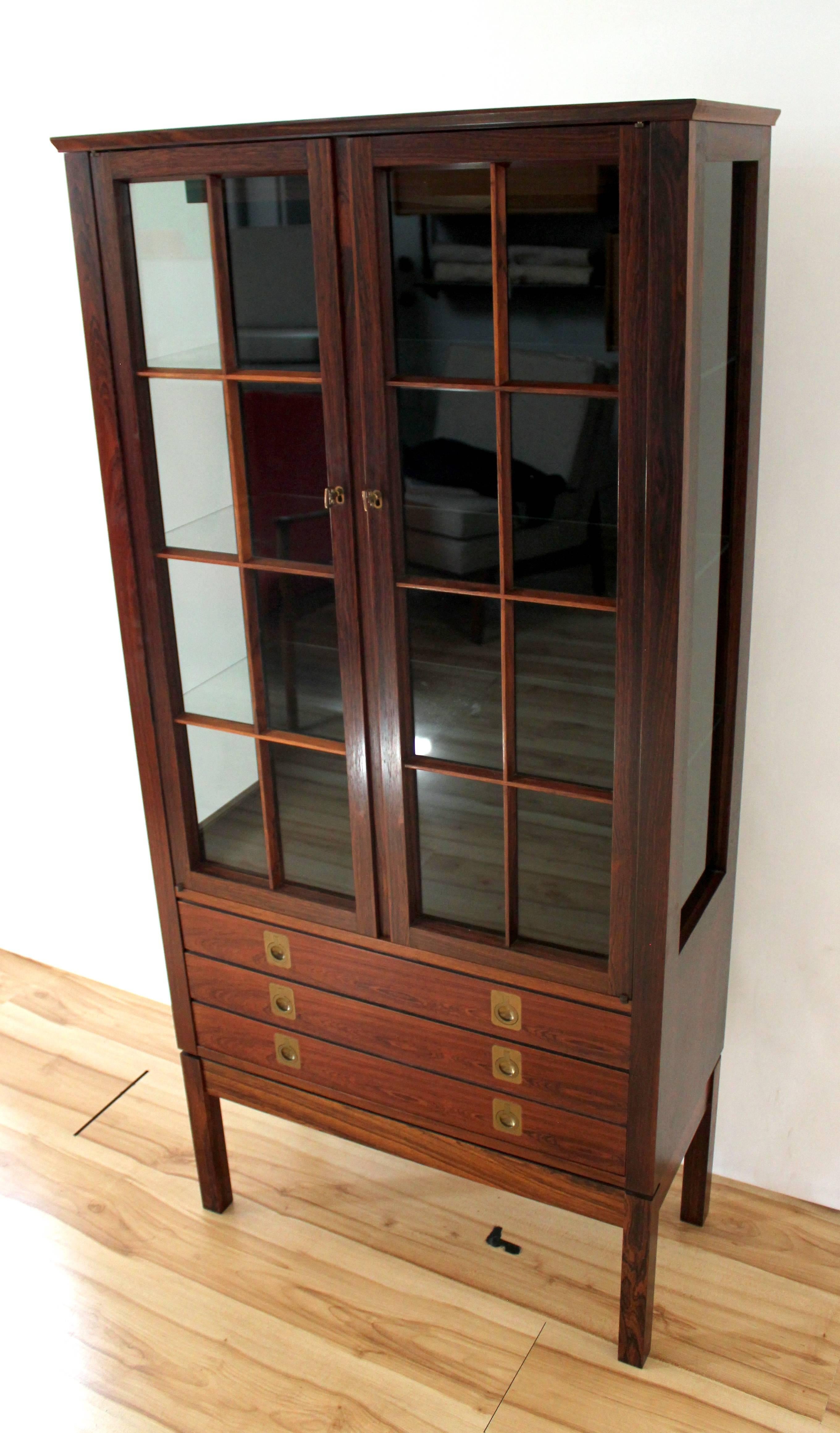 1960s rosewood lighted curio cabinet designed by Torbjørn Afdal for Bruksbo, made in Norway. Cabinet has two locking glass-fronted doors with keys, glass side panels, interior glass shelves, and three drawers with brass pulls.