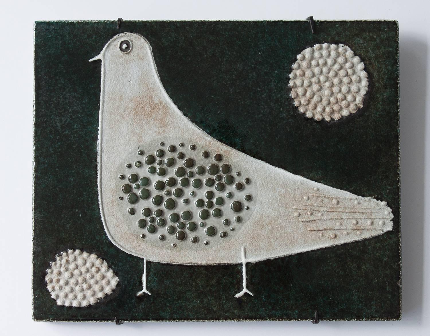 Decorative ceramic wall tile by Swedish artist Sylvia Leuchovius for Rorstrand. Stylized bird relief on dark blue-green glazed background. Original black iron mounting bracket. Sylvia's motifs are centered on plants, birds and children have been