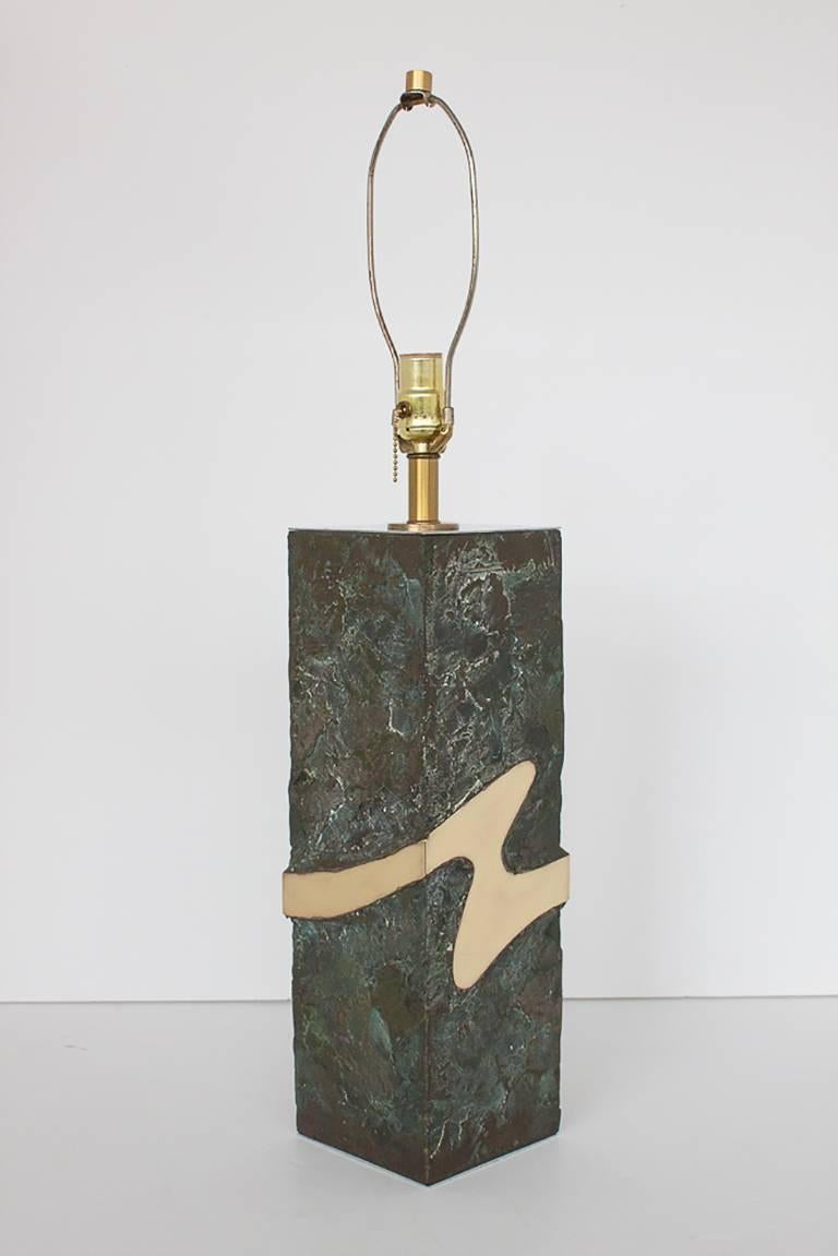 This mixed metal table lamp features an inlaid abstract solid polished brass form which wraps around all four sides of the lamp base. Realistic faux textured cast bronze surrounds the brass form. The textured bronze has a beautiful verdigris and