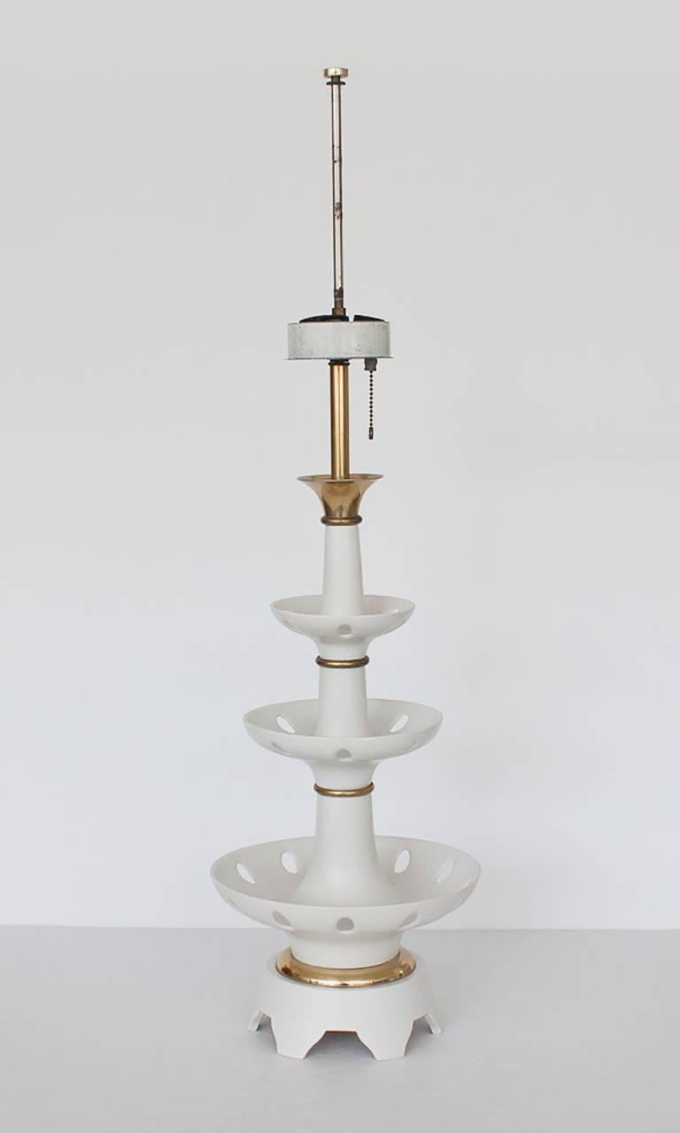 This table lamp features three-tiered / stepped white matte porcelain bowls with brass details between each segment. The bowls have pierced holes around the outside edge. Takes three standard light bulbs. Three-way pull chain. Solid brass finial.