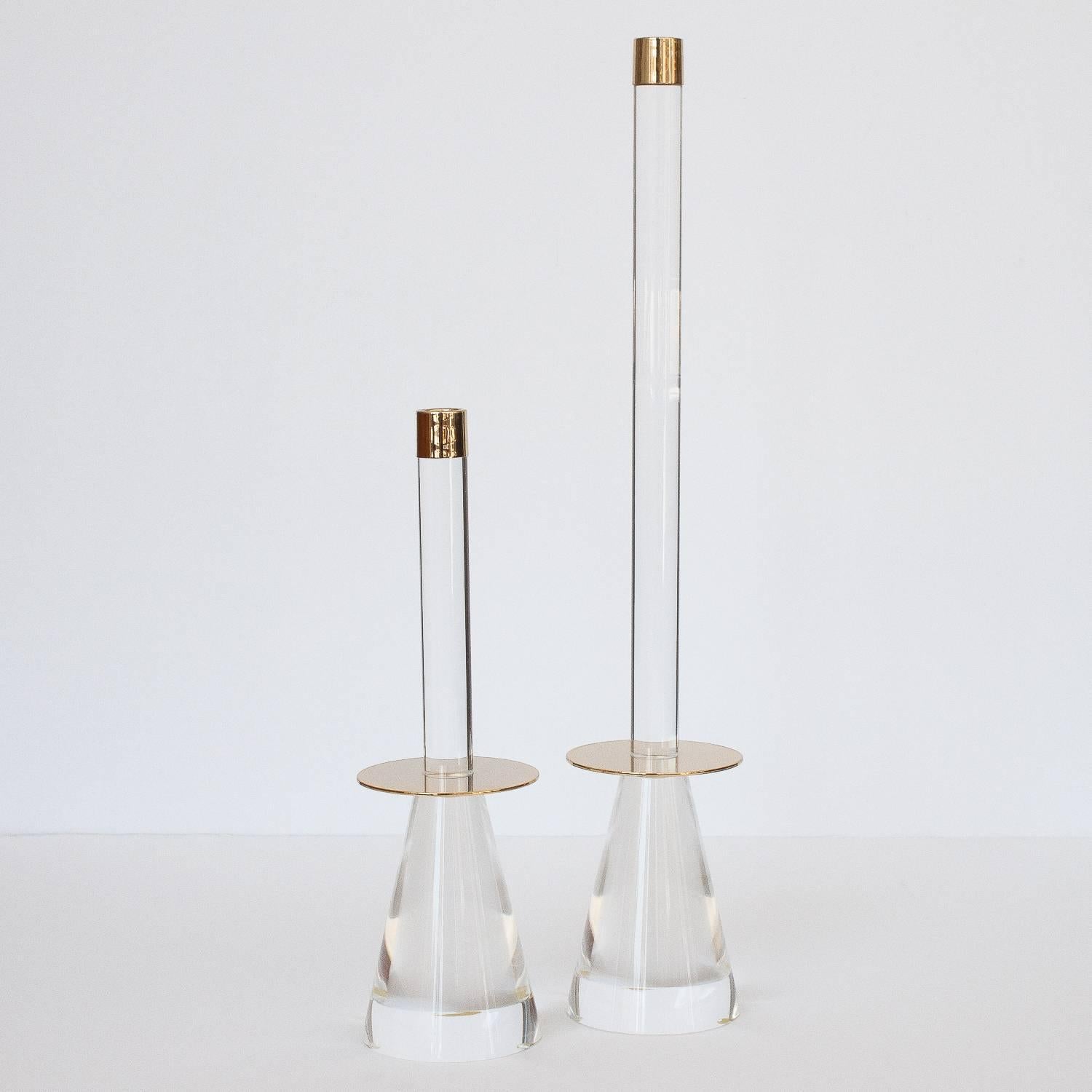 Pair of Lucite and brass candlesticks. Solid Lucite conical bases and stems. Polish brass discs separate the base and stem. Brass caps. Unmarked. 

Measures: Large 21.75" height x 4" diameter
Small 13.75" height x 4" diameter.