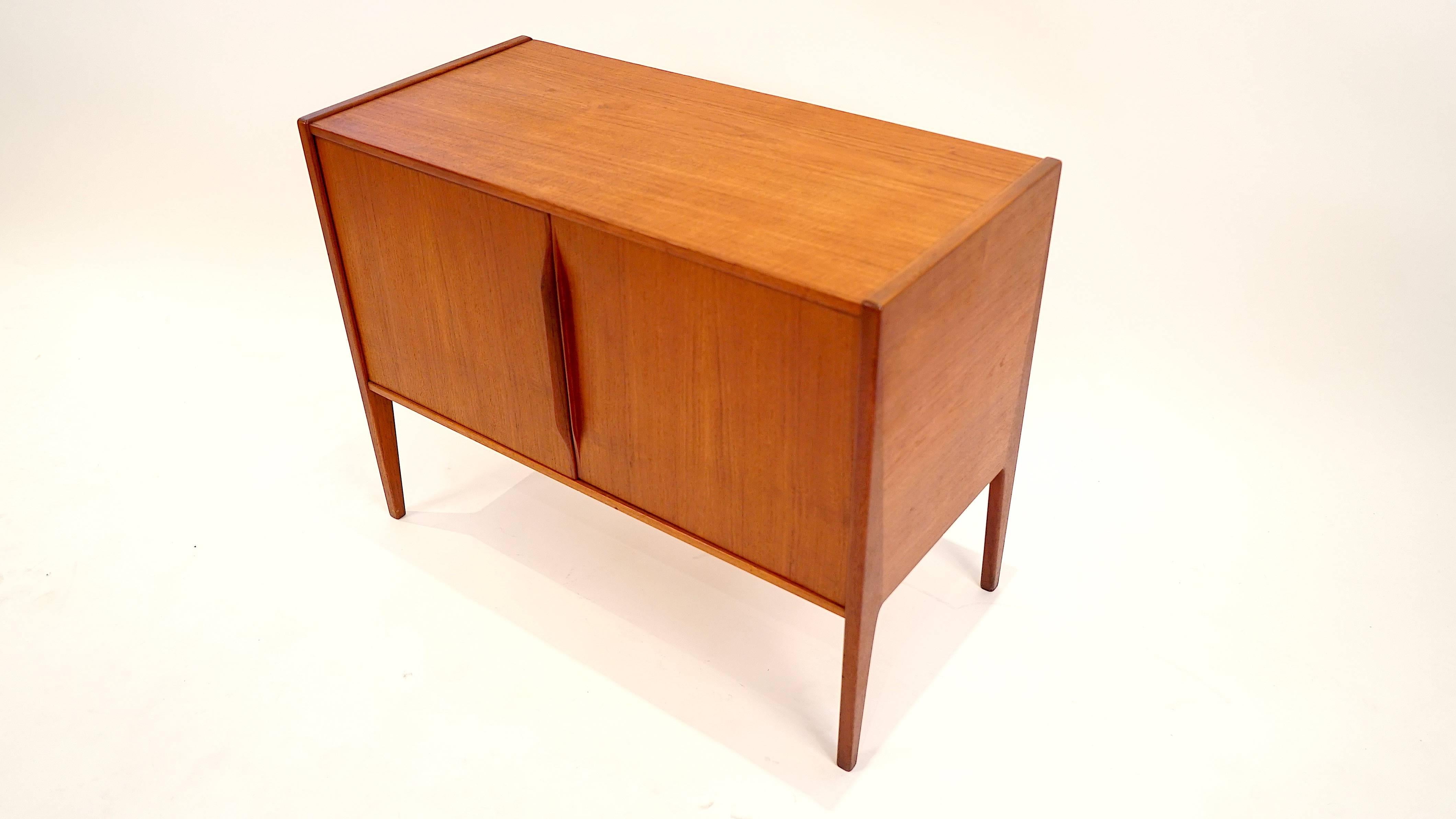Danish demi credenza with signature Nils Jonsson legs featuring two tone teak construction. Contoured door pulls accent the front and give way to storage space inside. Brass hardware and beautifully figured wood round out the design. This diminutive