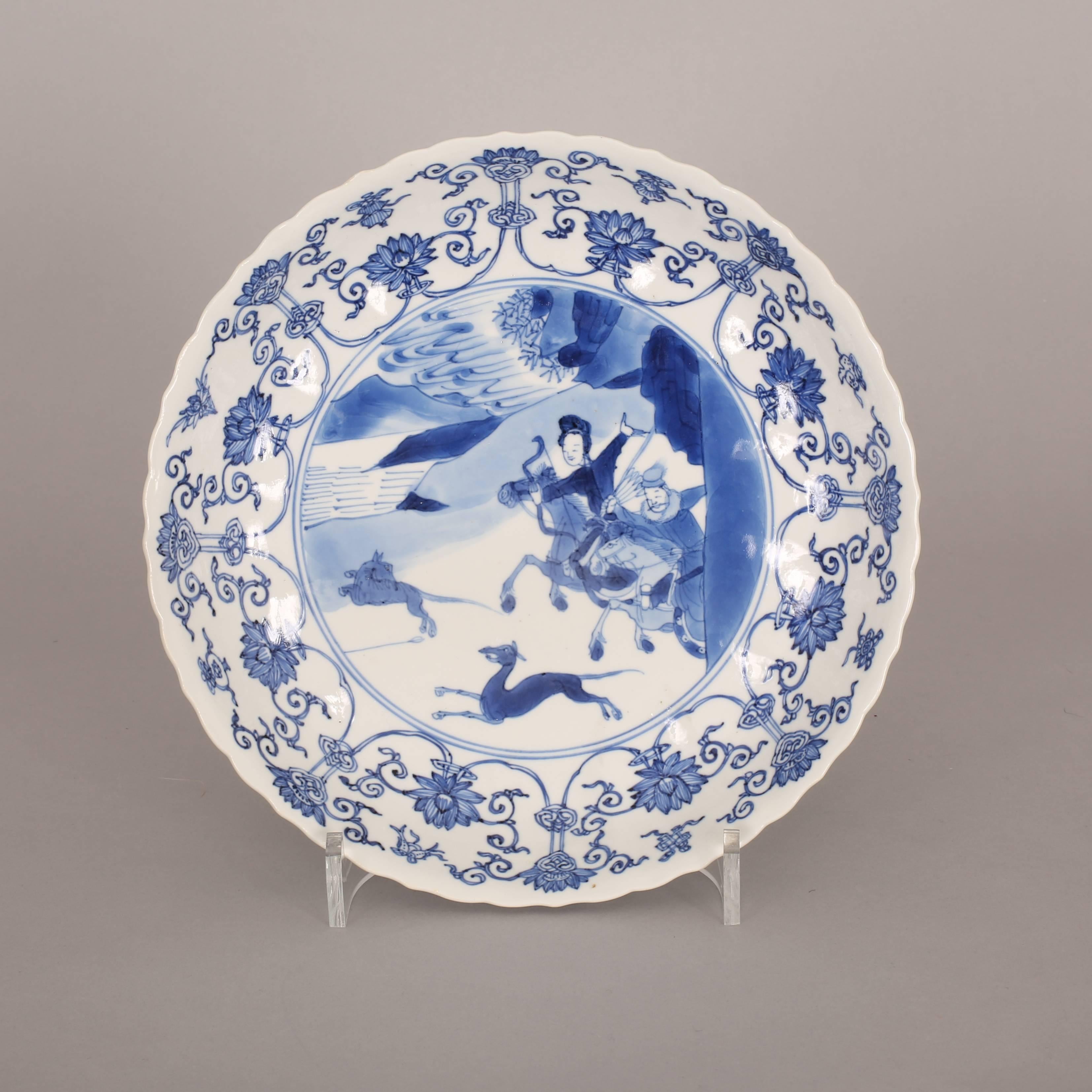 A Chinese porcelain blue and white saucer dish with foliate rim, painted with a central medallion of a hunting scene with a lady and a man both on horses with bows and arrows and two dogs in front, all in a mountainous river landscape scene, all