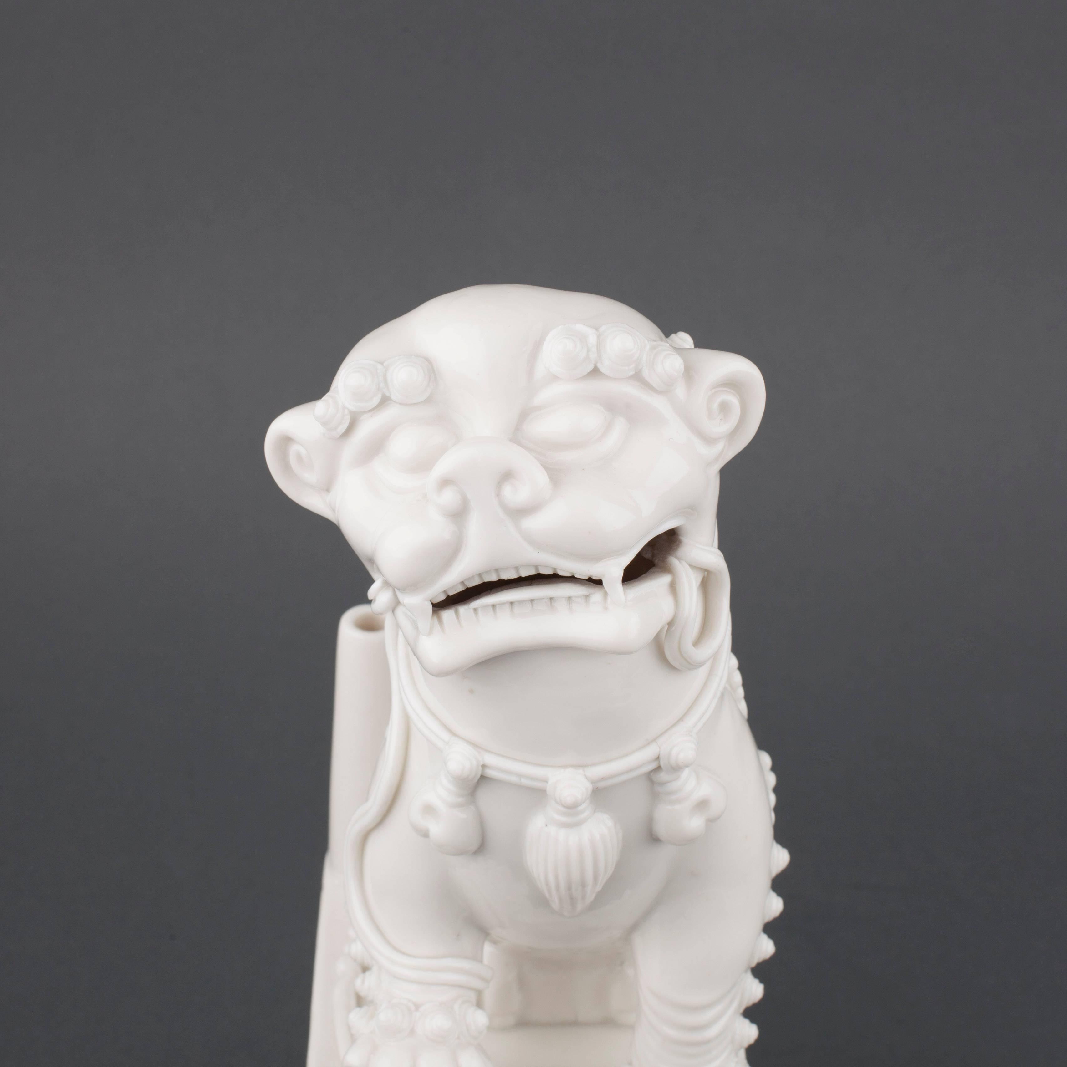 Chinese Blanc de Chine incense burner in the form of a seated Buddhist lion atop a square plinth, the head with open mouth, one paw resting on brocade ball, the neck tied with bells, decorated with a strap in his open mouth, with well moulded hair