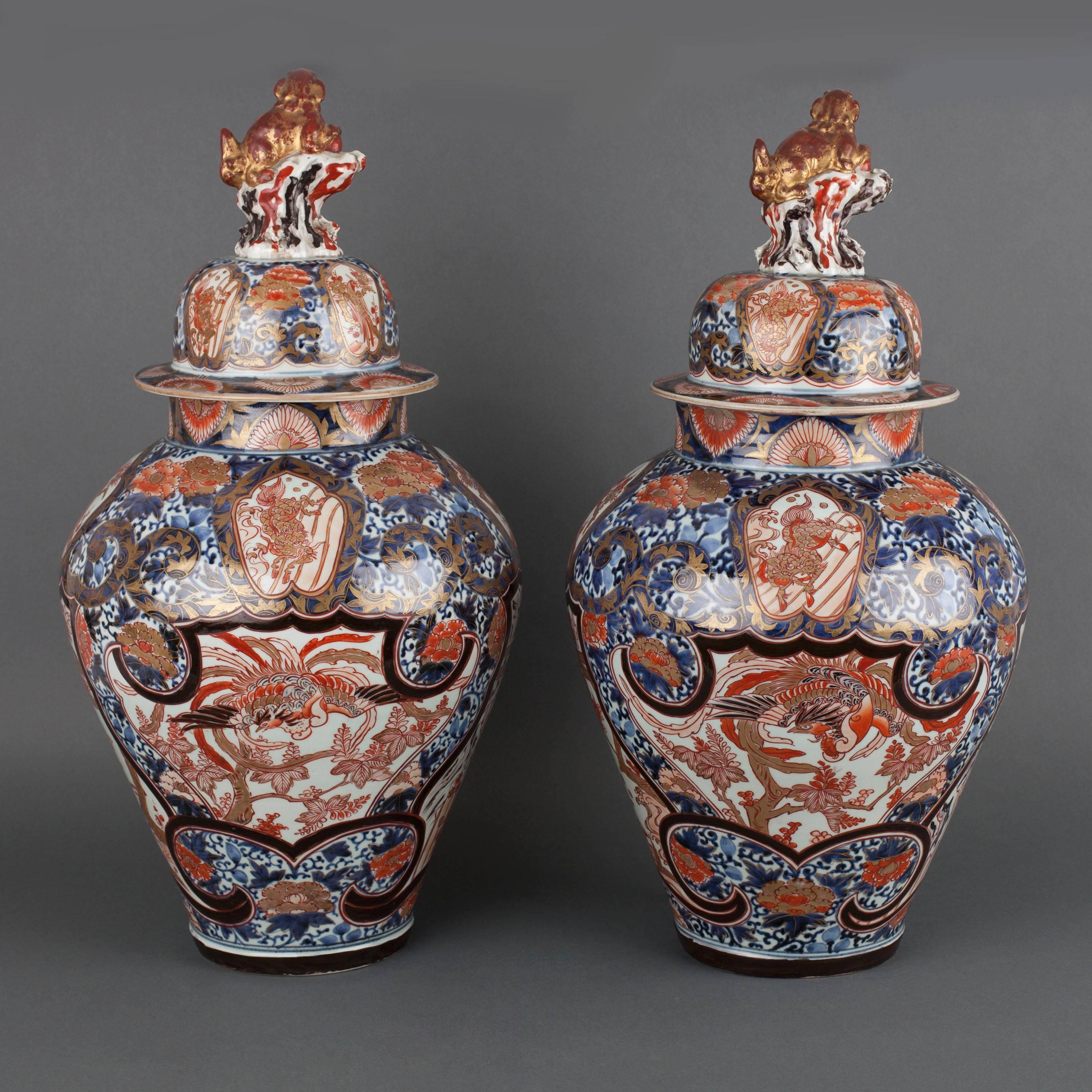 A pair of Japanese porcelain Arita Imari vases and covers decorated with iron-red phoenix birds within central panels with a black enamel border, the floral ground in underglaze-blue, the covers with Buddhist lion finials.

Measures: 60cm high