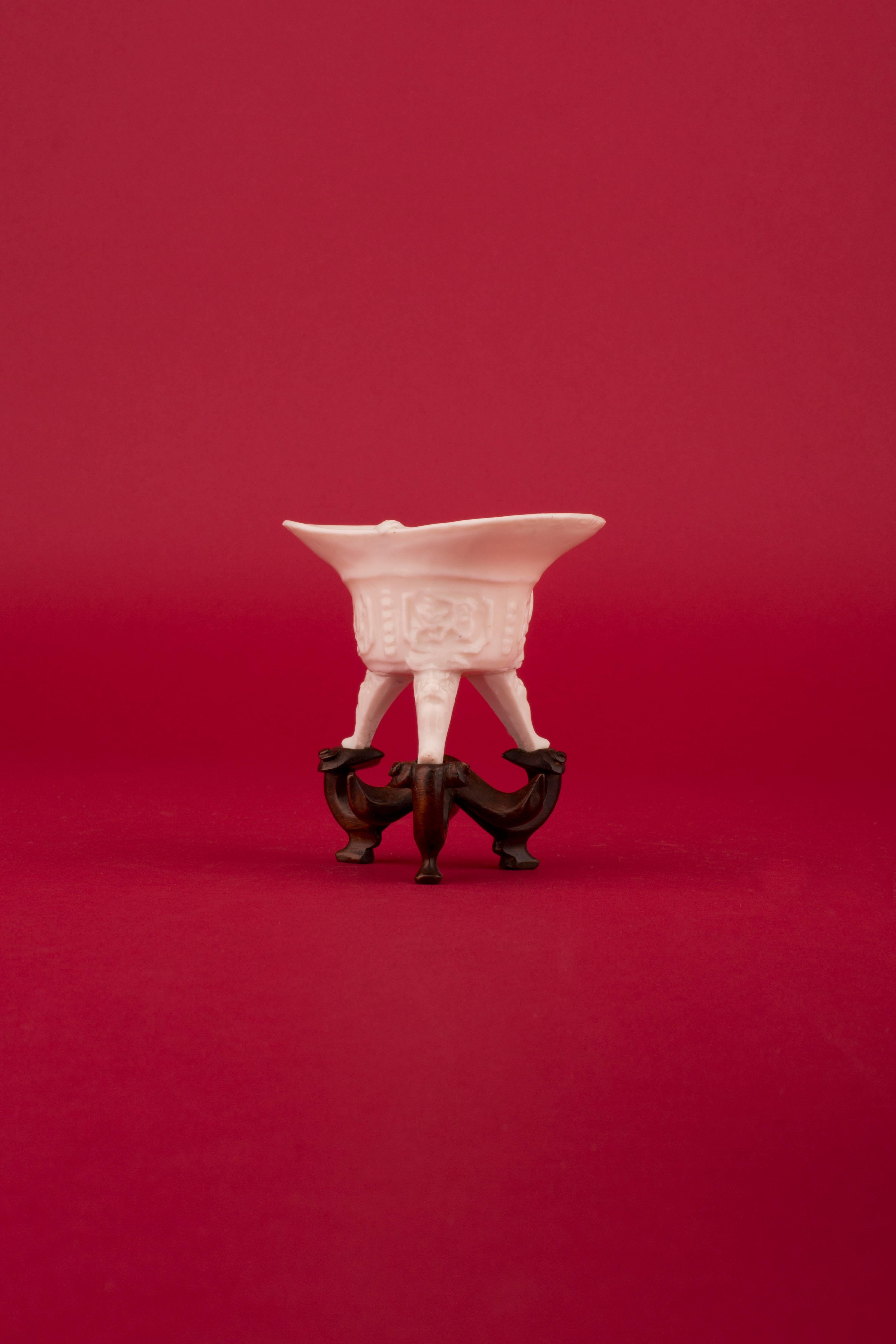 A Chinese porcelain blanc de chine jue, tripod libation cup with single strap handle, molded on the body with four panels of bamboo and lingzhi between relief angles, all on three feet with mask terminals and beneath a molded key-fret band at the