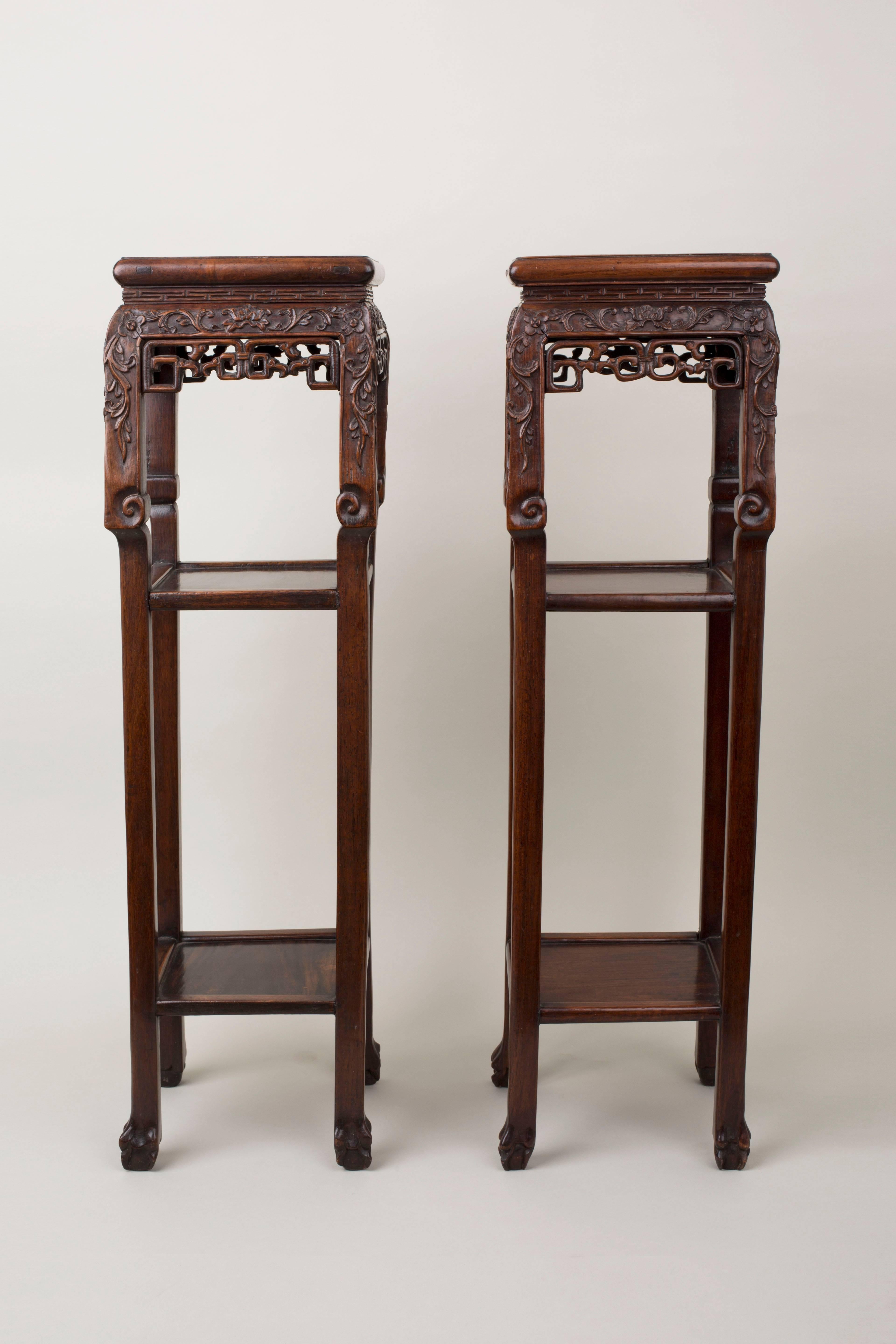 A pair of Chinese black wood hong mu square pedestal tables, the top inset with mottled red and white marble, the frieze carved in relief with flowers and scrolling foliage extending down the square legs which terminate above a low shelf into scroll