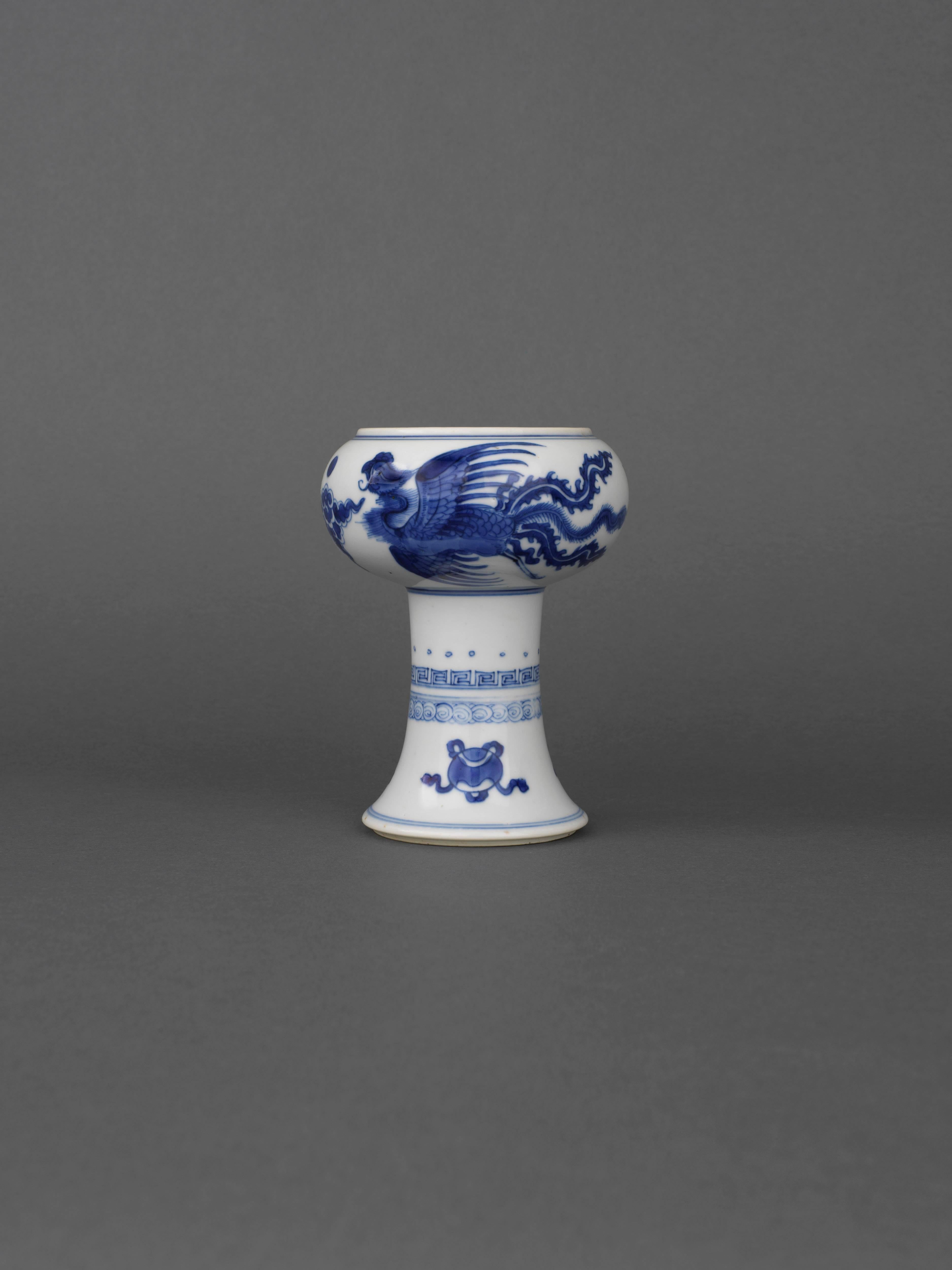 A Chinese porcelain underglaze blue and white stem cup painted on the body with two phoenix birds in flight between the moon and ruyi-head clouds between double rings, the stem with dot, key fret and scroll band, above ribbon tied pearl, lozenge and