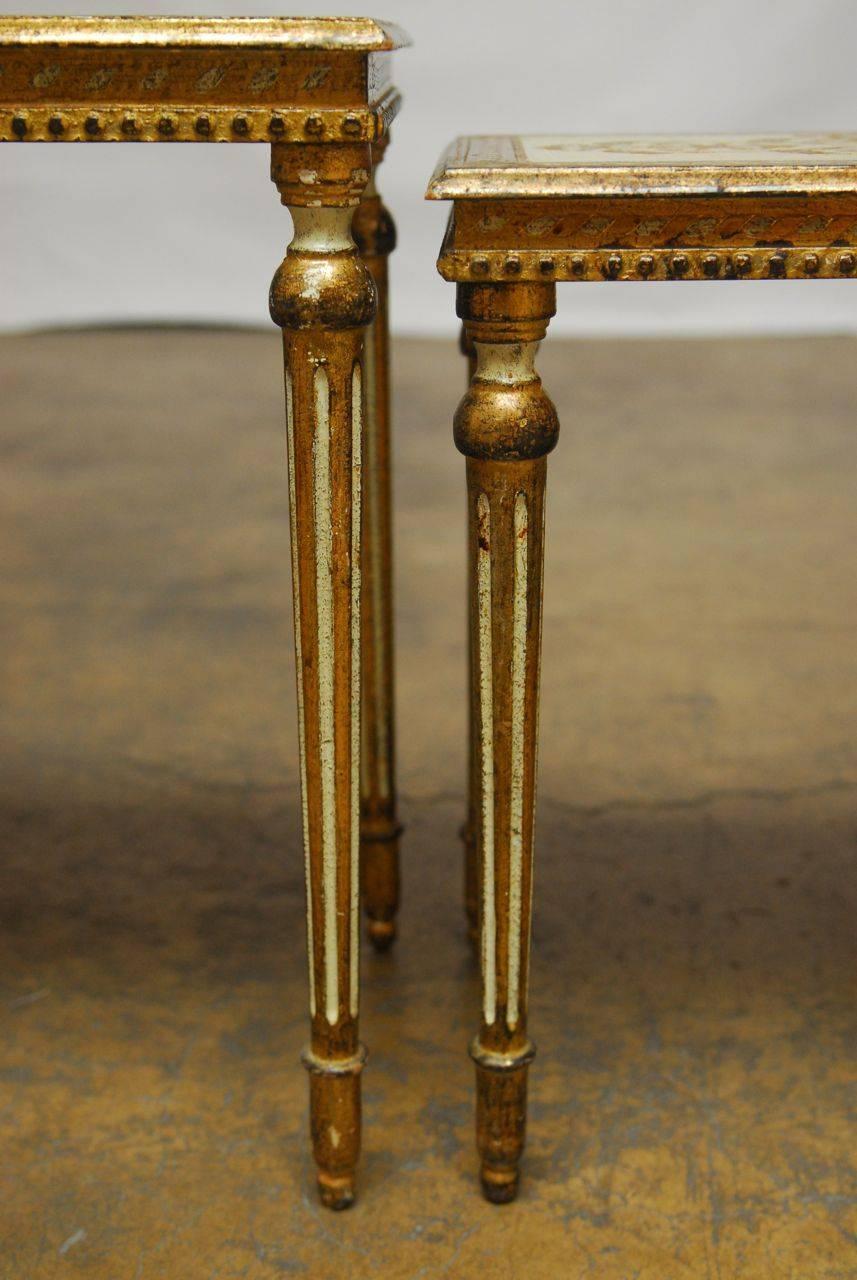 Set of lavish Italian nesting tables featuring Louis XVI style fluted legs and a hand applied gilt finish with a rich aged patina. Cream lacquer background on the tops with a foliate design and cream accents on the legs. Unique style, less common