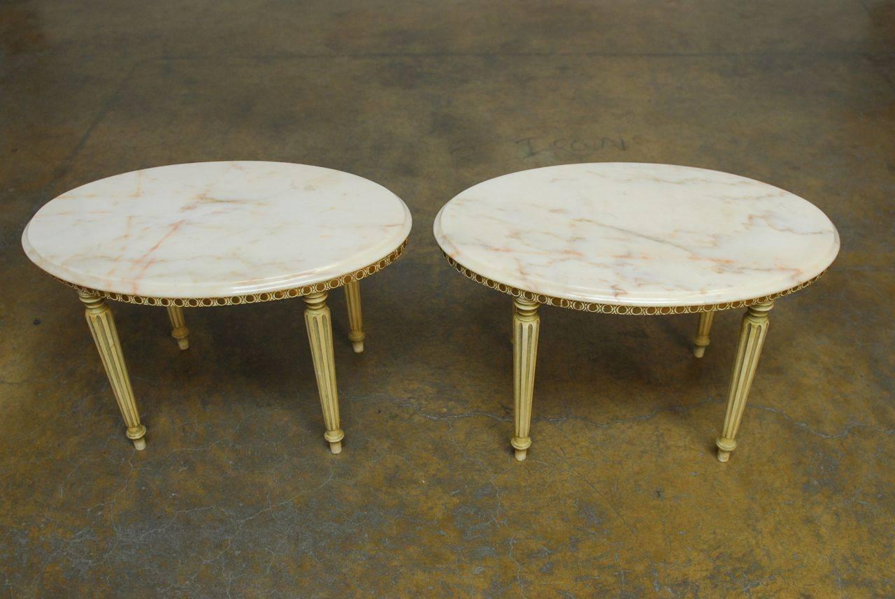 Elegant pair of drinks tables featuring oval marble tops each with an ogee edge. Made in the Louis XVI taste with Neoclassical styling and tapered, fluted legs that have been painted with a gilt apron and decorative edge. 