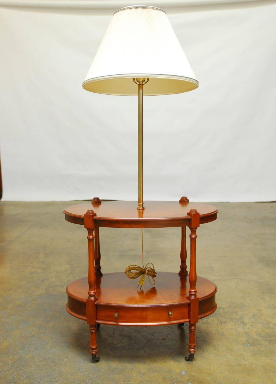 Oval two-tiered table featuring a built-in lamp supported by a fluted brass column by Frederick Cooper of Chicago. This lamp table has turned legs and sits on casters. The bottom shelf has a storage drawer with brass pulls. The top tier is 22
