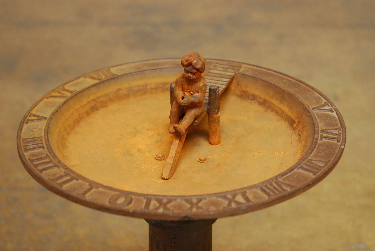 Solid cast iron English garden birdbath featuring a sundial around the side of the bowl and a figural child sitting on a pier. Heavy and patinated with a vintage, weathered patina.