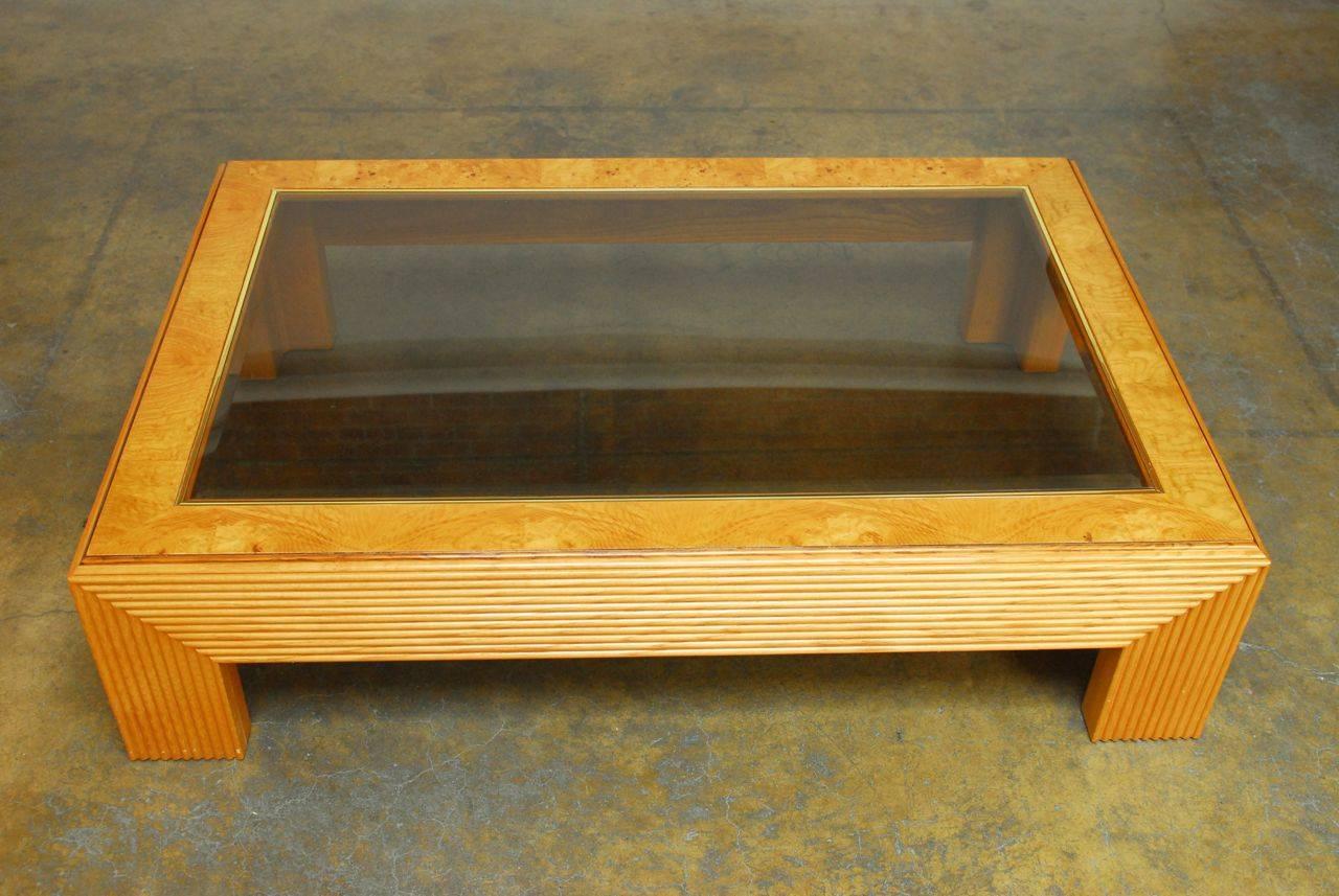 Large coffee table influenced by the architecture of the streamlined Art Deco period. Geometric structural style legs and sides draw inspiration from a skyscraper skyline. The top features a burl wood veneer and a beveled, tinted glass with a brass