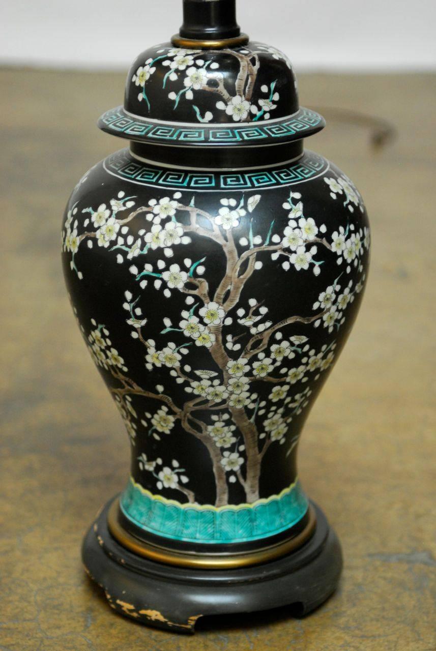 Chinese famille noir style ginger jar mounted as a table lamp featuring a prunus blossom motif, accented by ringed Greek keys. Stunning white prunus blossoms on the tree stand out against the black background with highlights of teal and yellow.