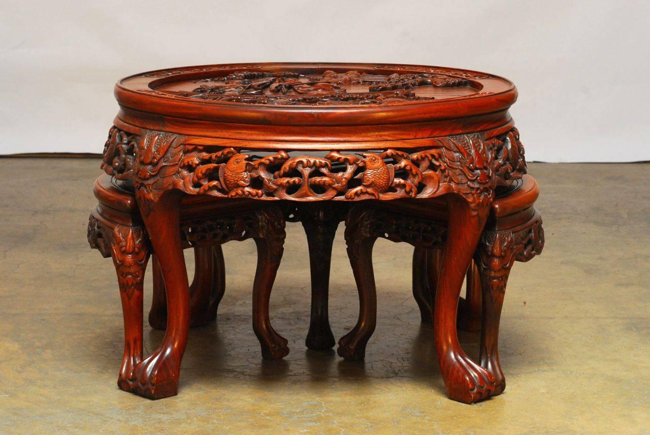 Elegantly designed tea table with four stools nested below in a conforming shape. Table and stools feature deep, ornate relief carvings of dragons and koi throughout the aprons and legs. Tabletop features a carving of two warrior women with swords