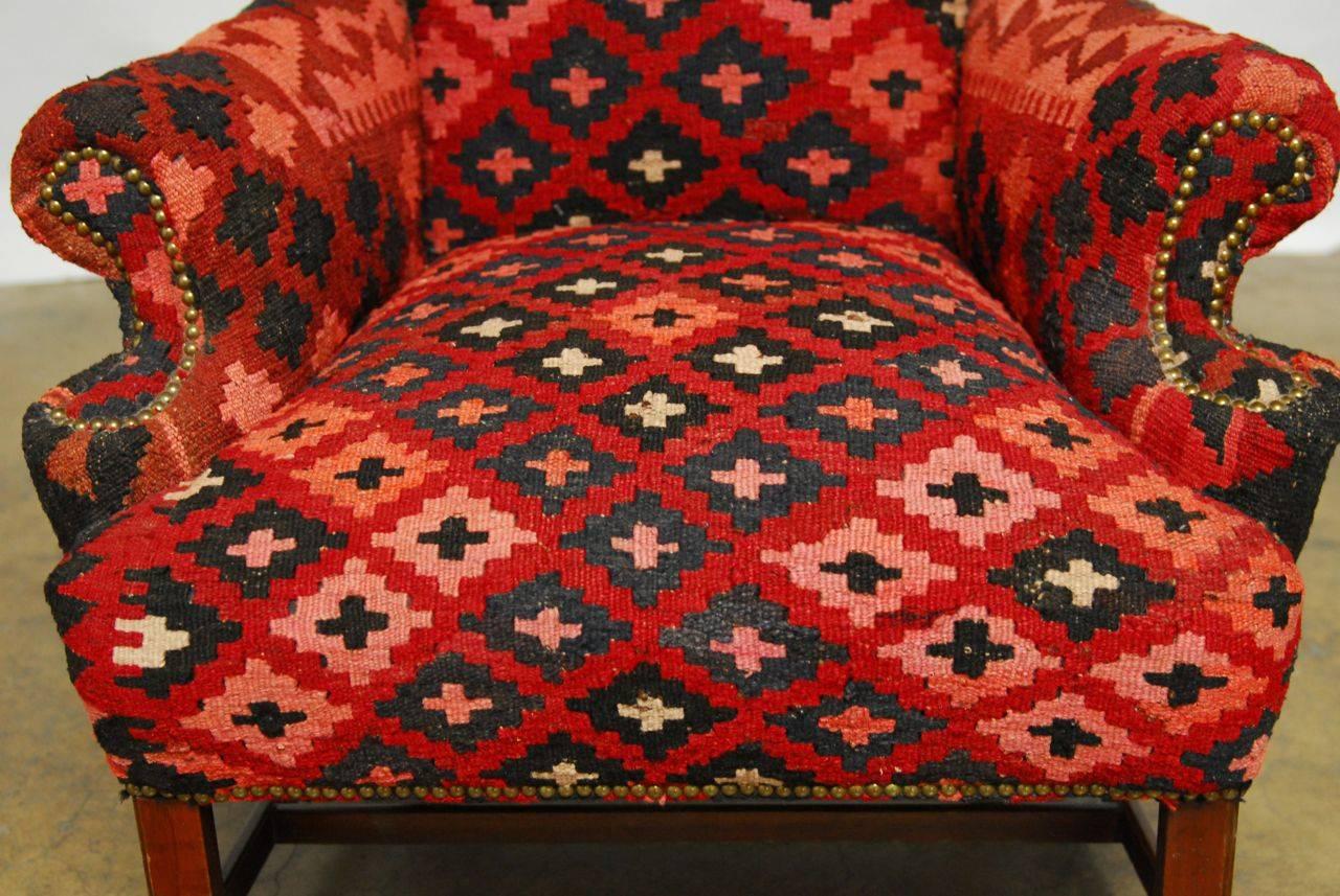 Fine wing chair upholstered in a handwoven geometric kilim rug made in a tribal style. This carpet features shades of red and black with pink accents. Brass nailhead trim combine to create a visually stunning and chic chair. Generous size with fully