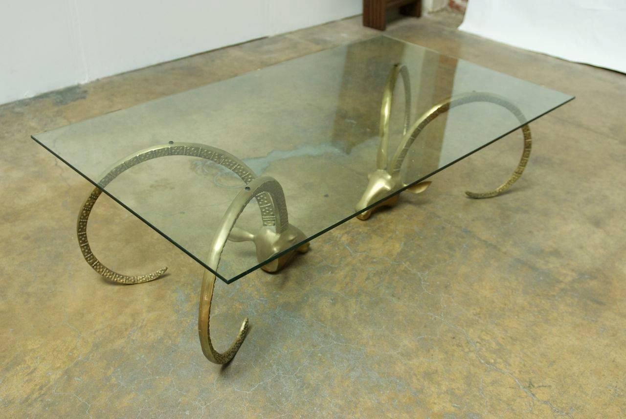 Impressive glass coffee cocktail table featuring a pair of brass Ibex or ram’s head bases with long patterned horns. Fabulous Hollywood Regency style from a local estate who purchased them in Carmel California, 1975.