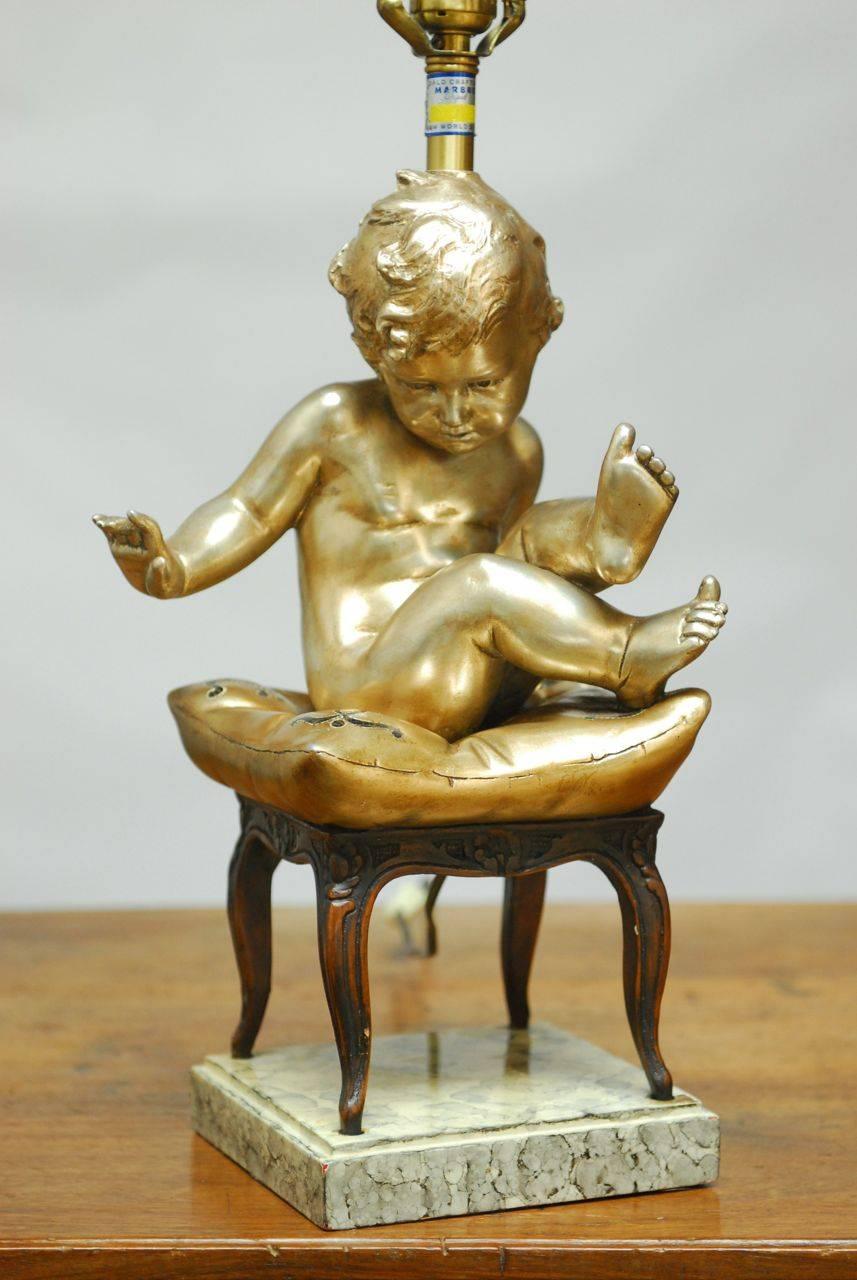 Stunning table lamp featuring a silver gilt Putti sitting on a gilt pillow and perched upon a carved French table with cabriole legs. This rare lamp by Marbro company is made of plaster and wood with brass hardware and rests on a faux marble
