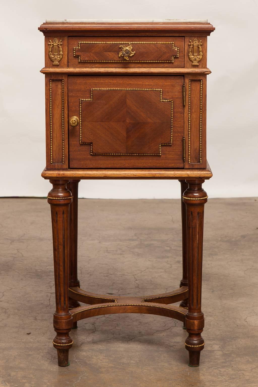 Ornate nightstand with a marble top featuring ormolu-mounted lyre shaped embellishments and trim. Lovely veneered case fronted by one drawer and one door below with a porcelain lined storage area. Supported by tapered and fluted legs with curved