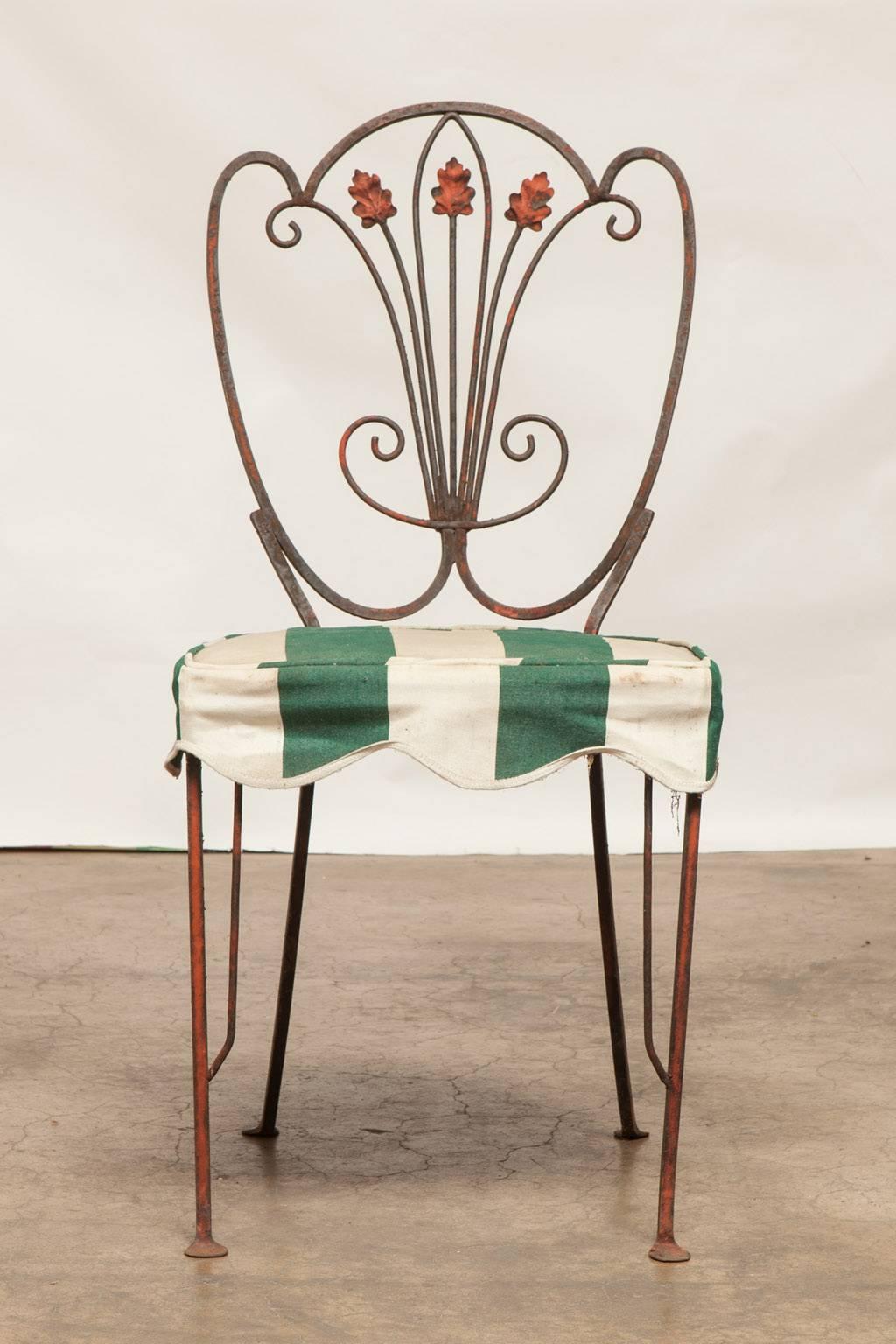 Charming set of French bistro dining chairs featuring a heart shaped back with a floral scrolled back. Wooden chair pads with vintage covers in a green and white cabana stripe. The finish on the chairs is well patinated and three chairs are missing