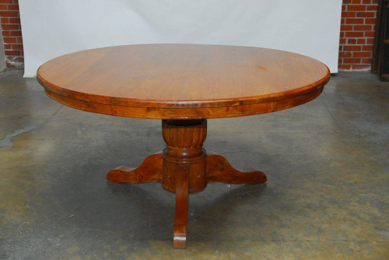 Grand, solid teakwood pedestal dining table hand-carved with a large round top supported by a thick urn-shaped pedestal and three feet. Suitable for outdoor use with a rich grain pattern and quality finish. Made in the colonial style. This table