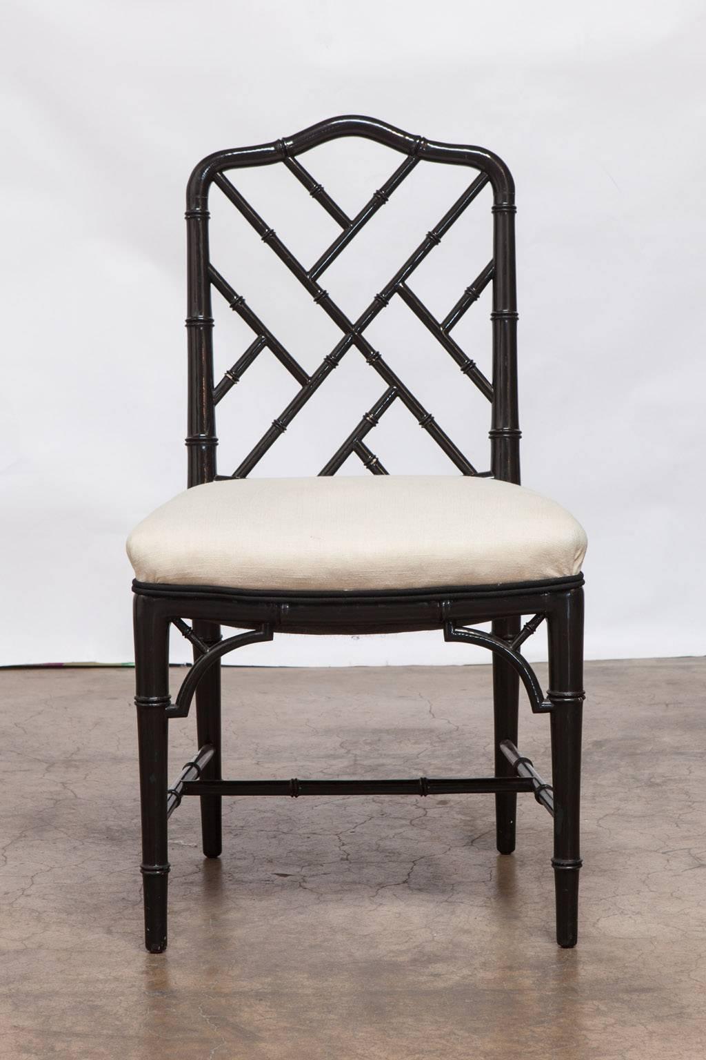 Stylish set of four faux bamboo dining chairs made in the Chinese Chippendale taste featuring a black lacquer finish. Upholstered in a cream color linen with a black double welt. Intricately carved frames with an open fretwork back and decorative
