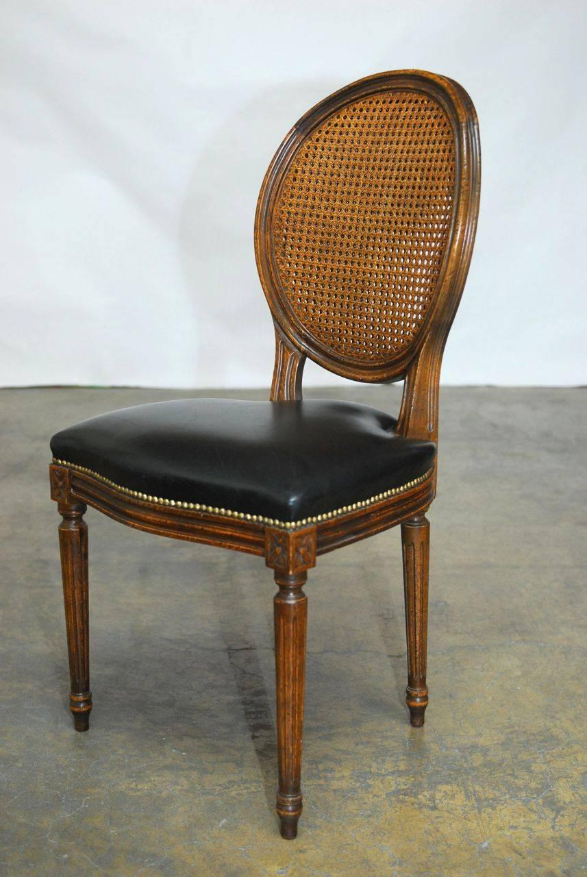 Lavish set of four Louis XVI style dining chairs featuring a double cane back and a black leather seat upholstery accented by brass nailhead trim. Exquisitely carved frames with a serpentine seat and carved rosettes on the molded frame. Supported by