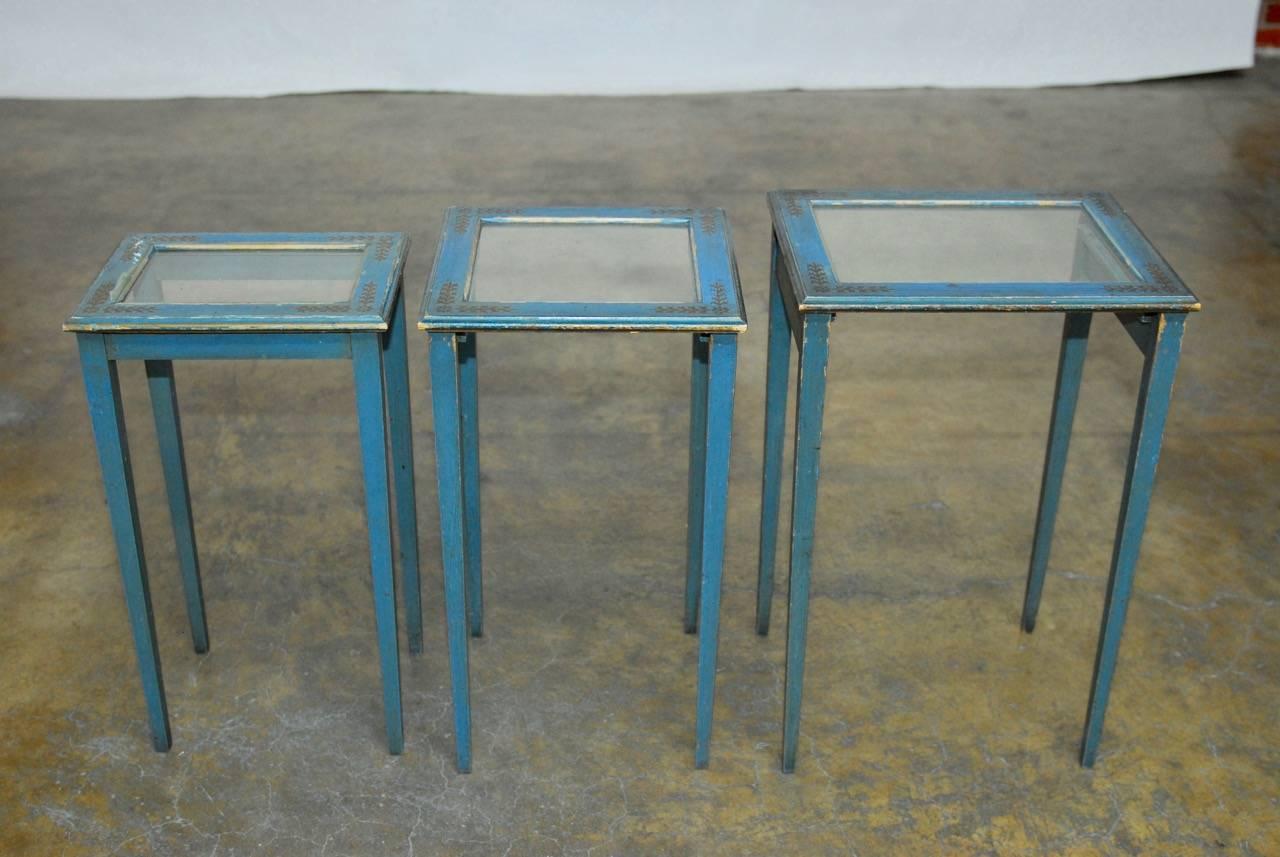 Unique set of nesting tables painted in a French blue with laurel motif gilt decoration. Attributed to Peter Engel Inc., New York. These tables have a framed glass top and slide together. Supported by delicate tapered legs with an aged vintage