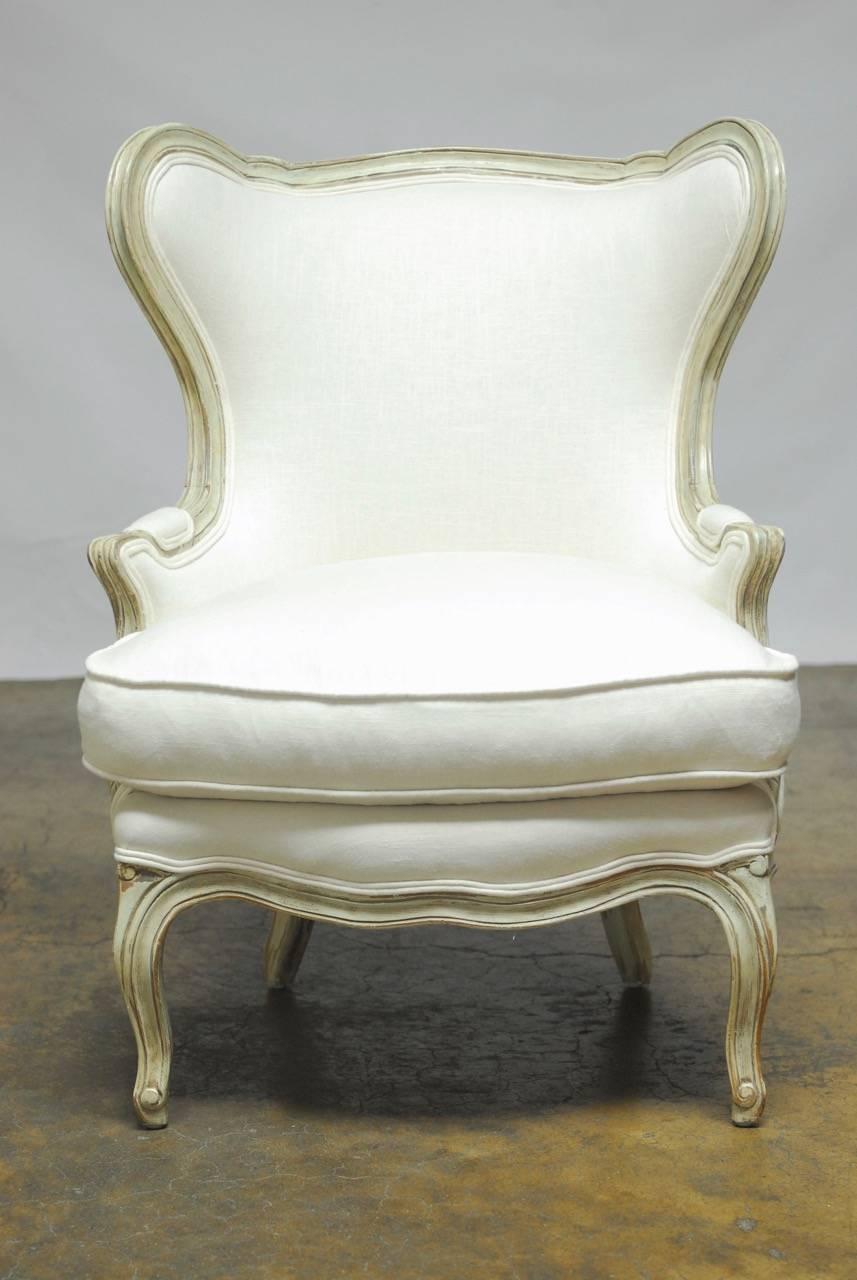 Spectacular wingback chair and ottoman newly upholstered in cream Irish linen featuring a painted frame made with generous proportions. Hand-carved in the Louis XV taste but with a Mid-Century Modern style wide wing butterfly chair design. The